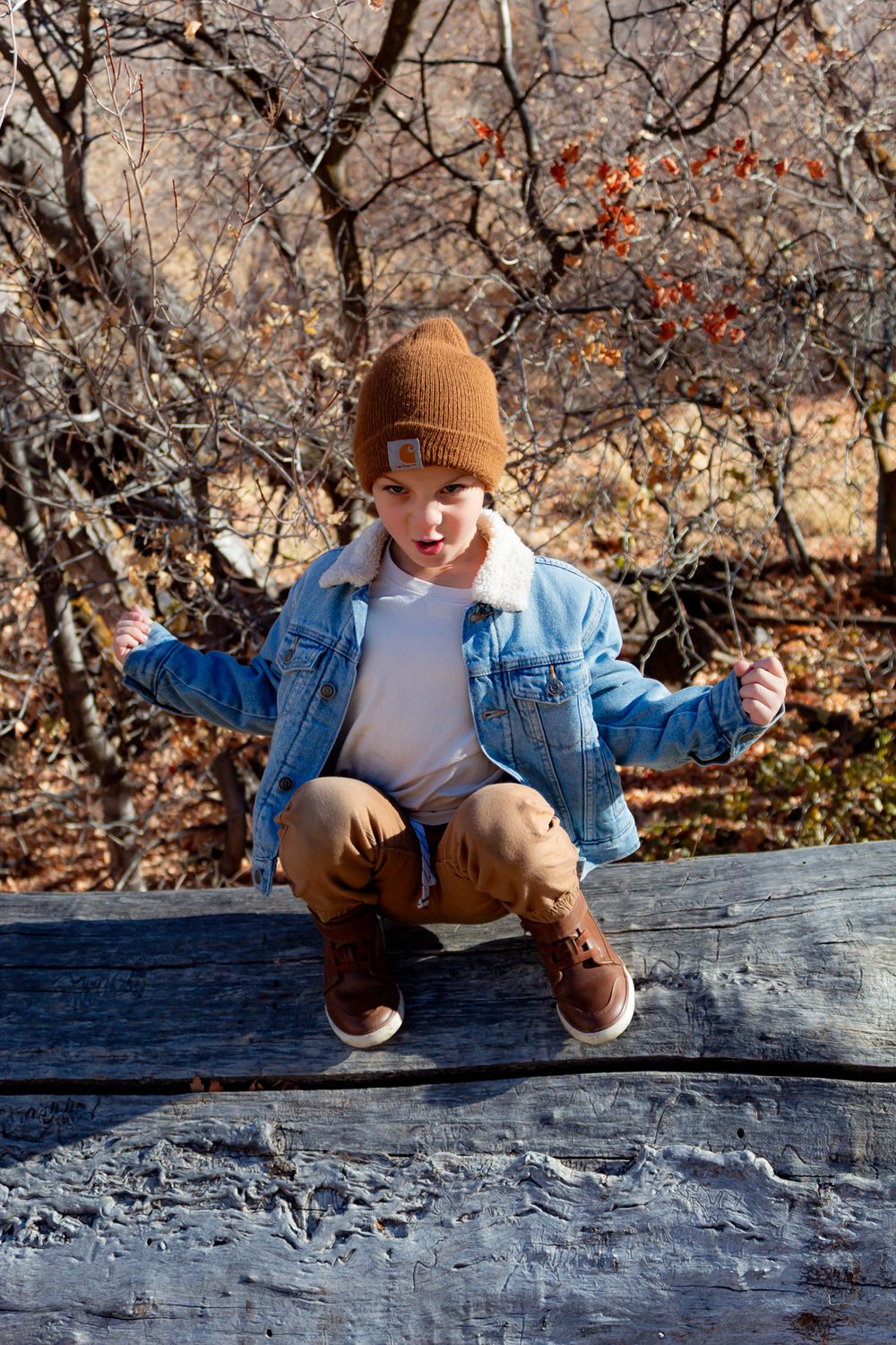 Person in denim jacket and beanie sits casually on fallen log in autumn forest.