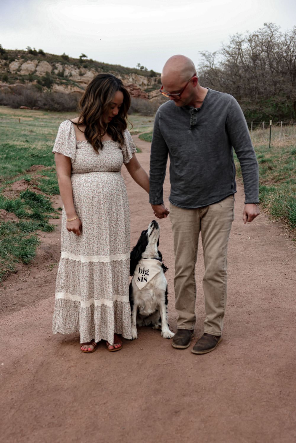 Happy couple walking on a dirt trail with their dog against scenic rocky backdrop and fields.