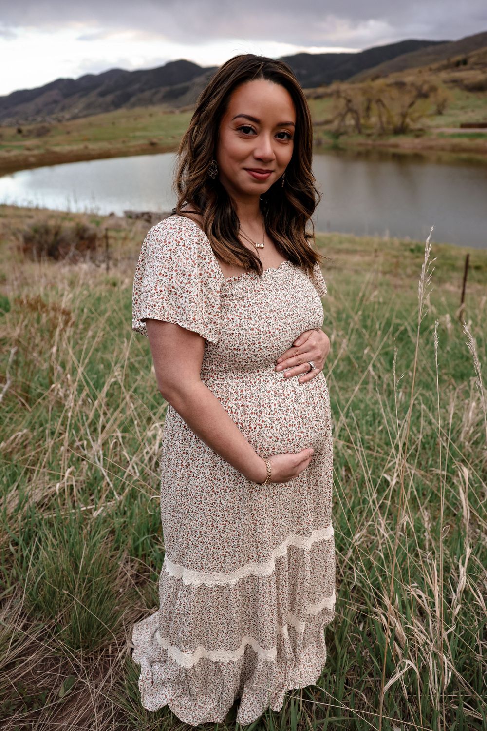 Woman in floral maxi dress poses in front of mountain lake landscape.
