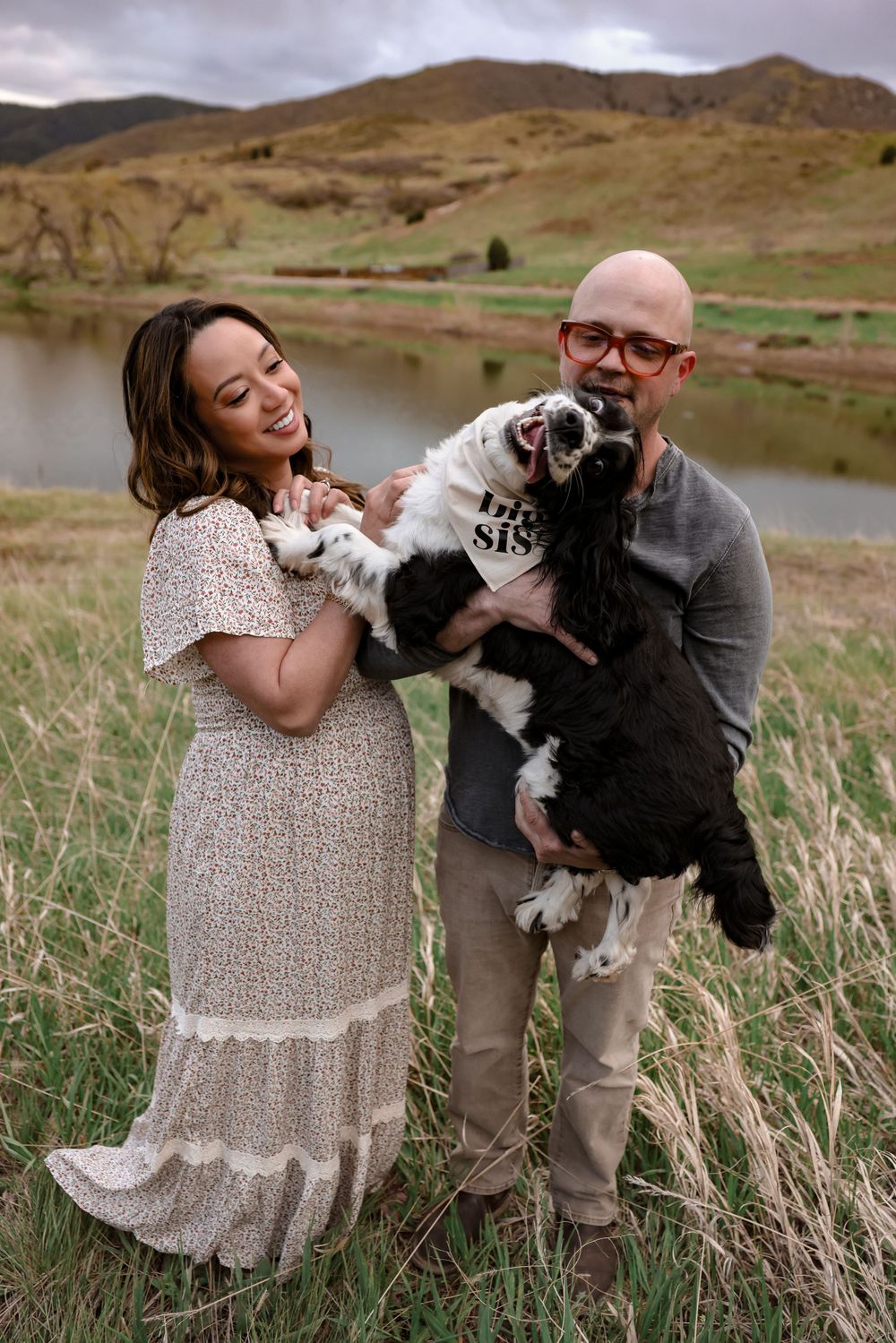 Couple holding their black and white dog while standing by mountain lake.
