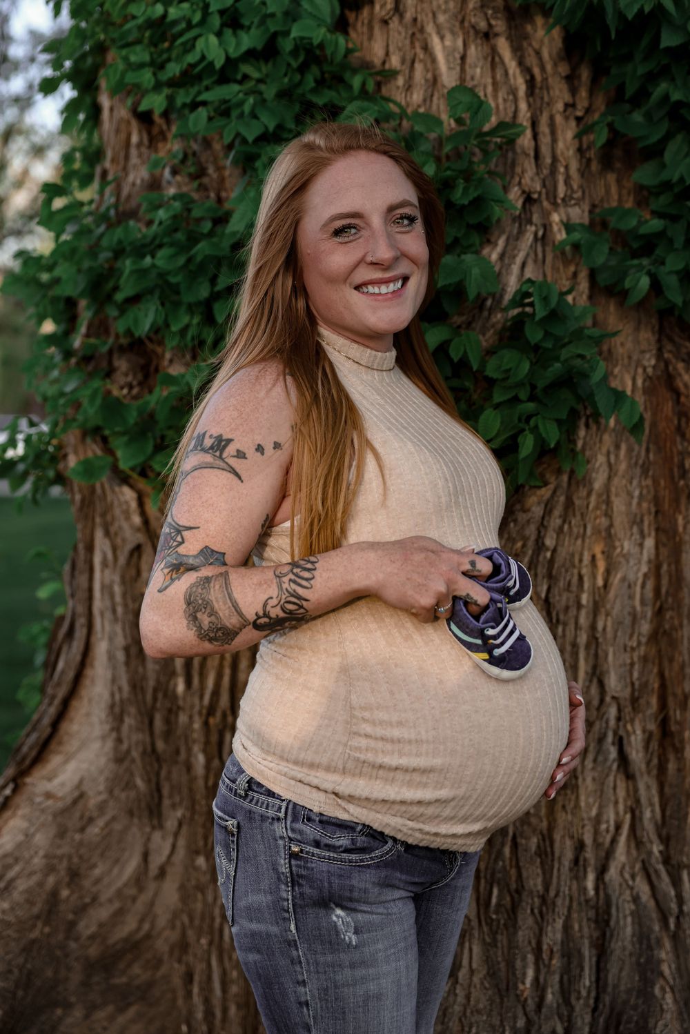 Expectant mother in beige sweater shows tattoos while holding baby shoes by tree.