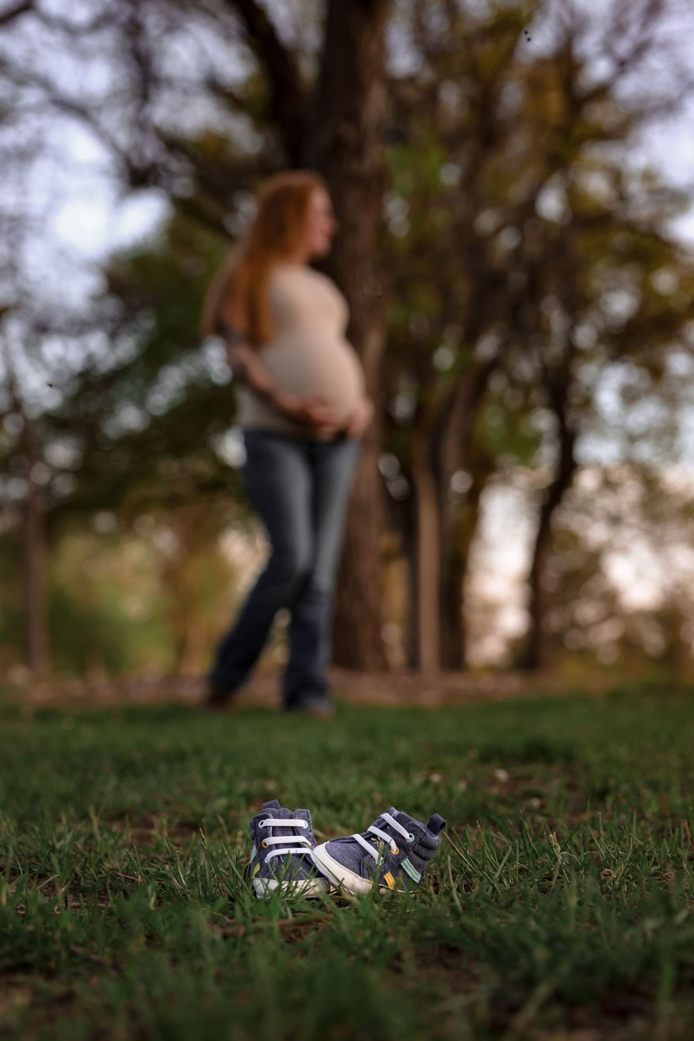 Baby shoes in focus on grass with expectant mother blurred in background.