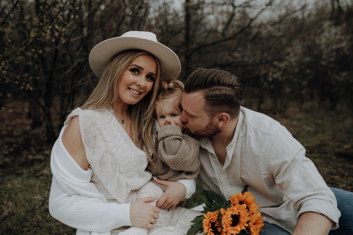 Family portrait with smiling mother in hat, father kissing toddler holding sunflowers outdoors.