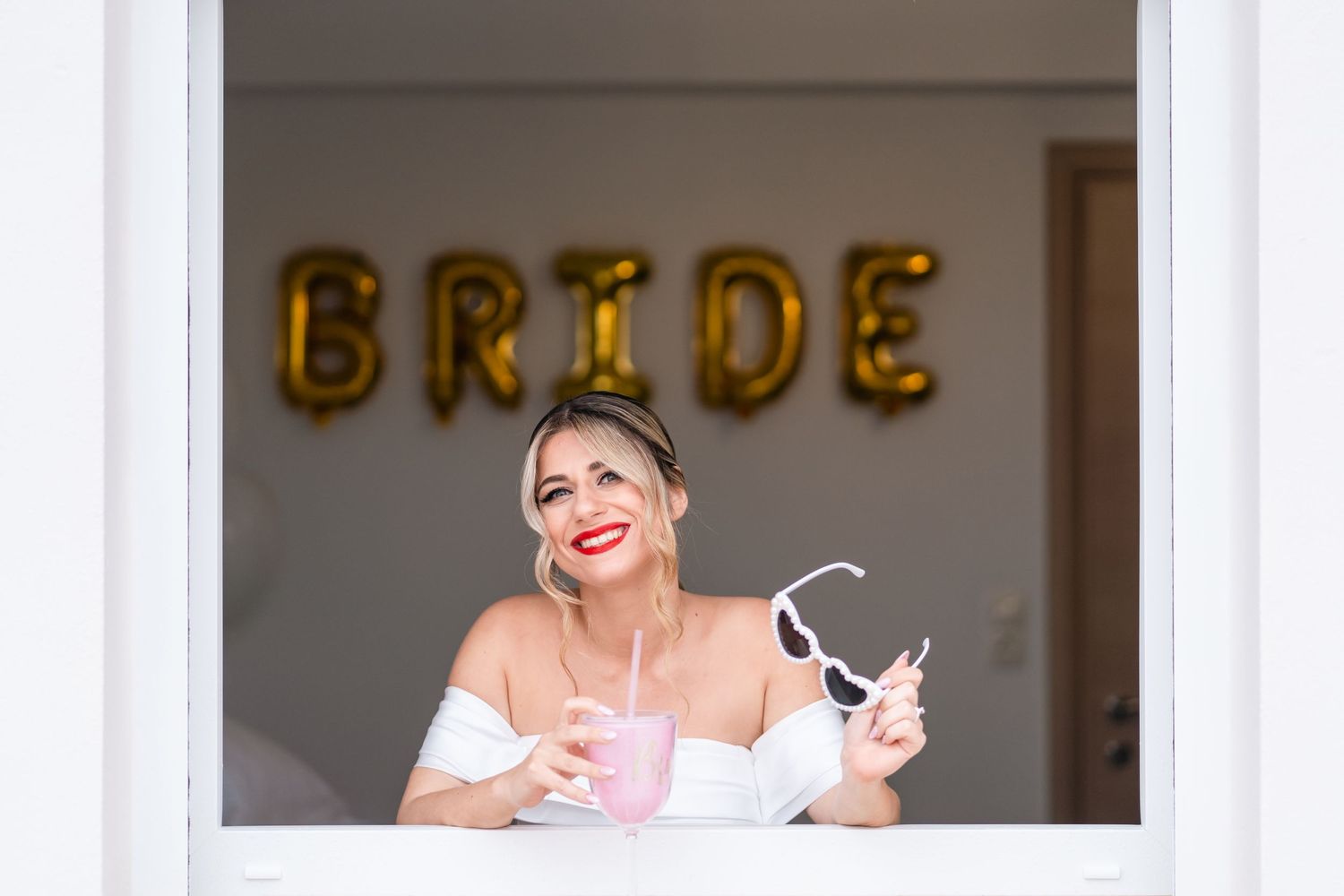 A bride smiles through a window with gold 'BRIDE' balloons visible in the background.