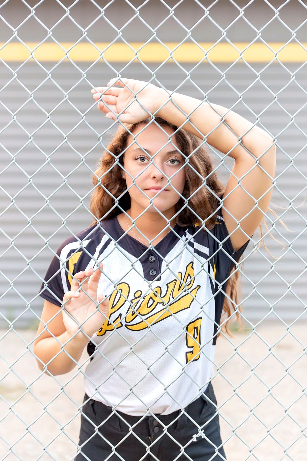 Softball player in Lions uniform poses behind chain link fence.