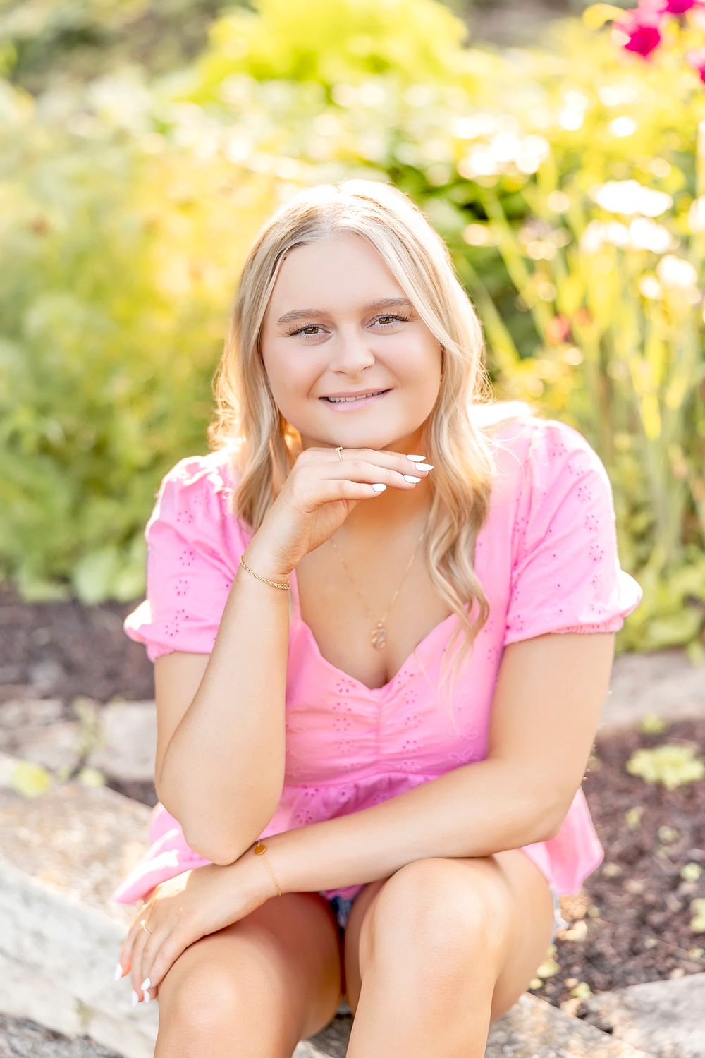 Woman in pink dress poses in garden setting with flowers during sunset.