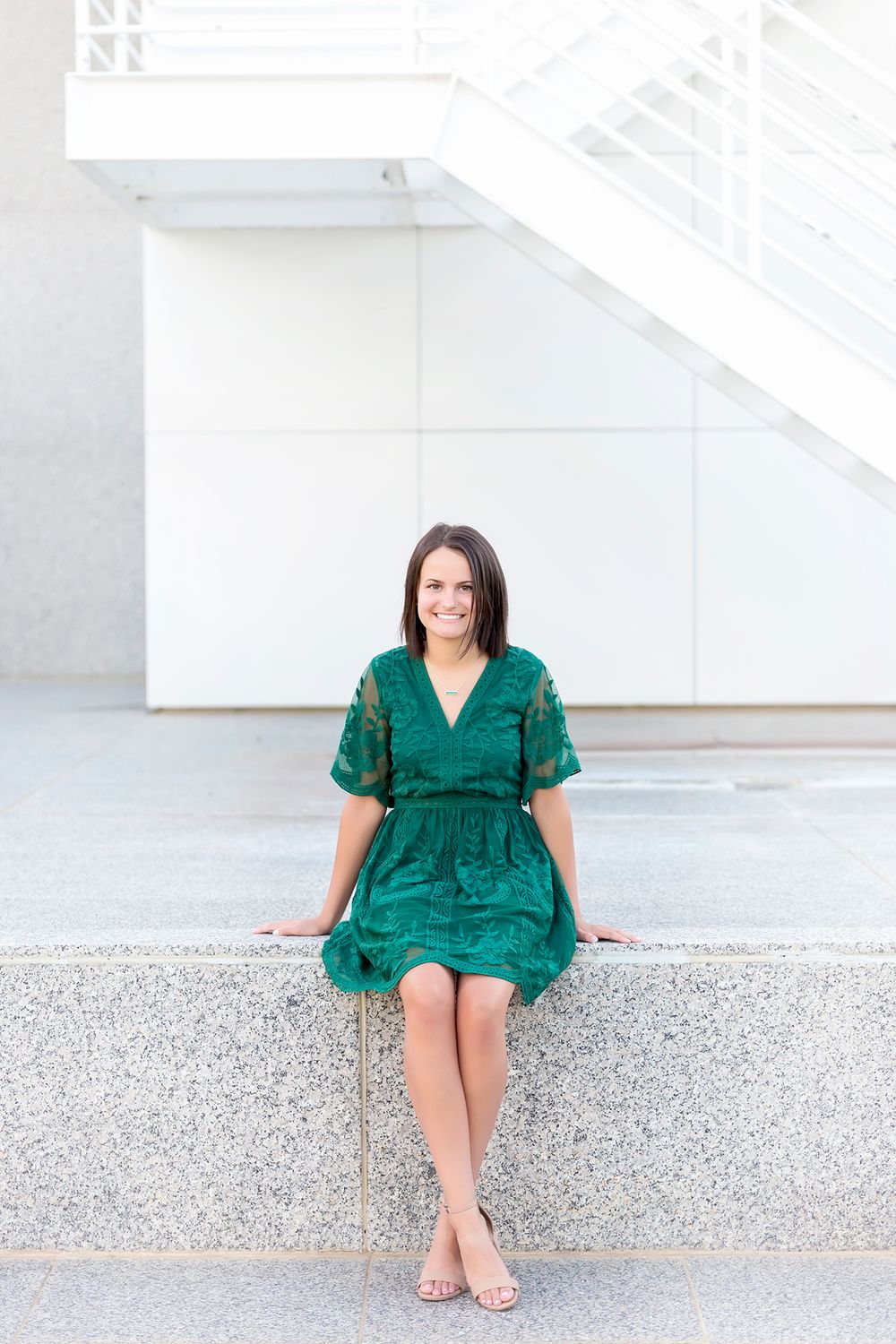 A woman in a vibrant emerald green dress poses outdoors on a concrete ledge in multiple casual positions.