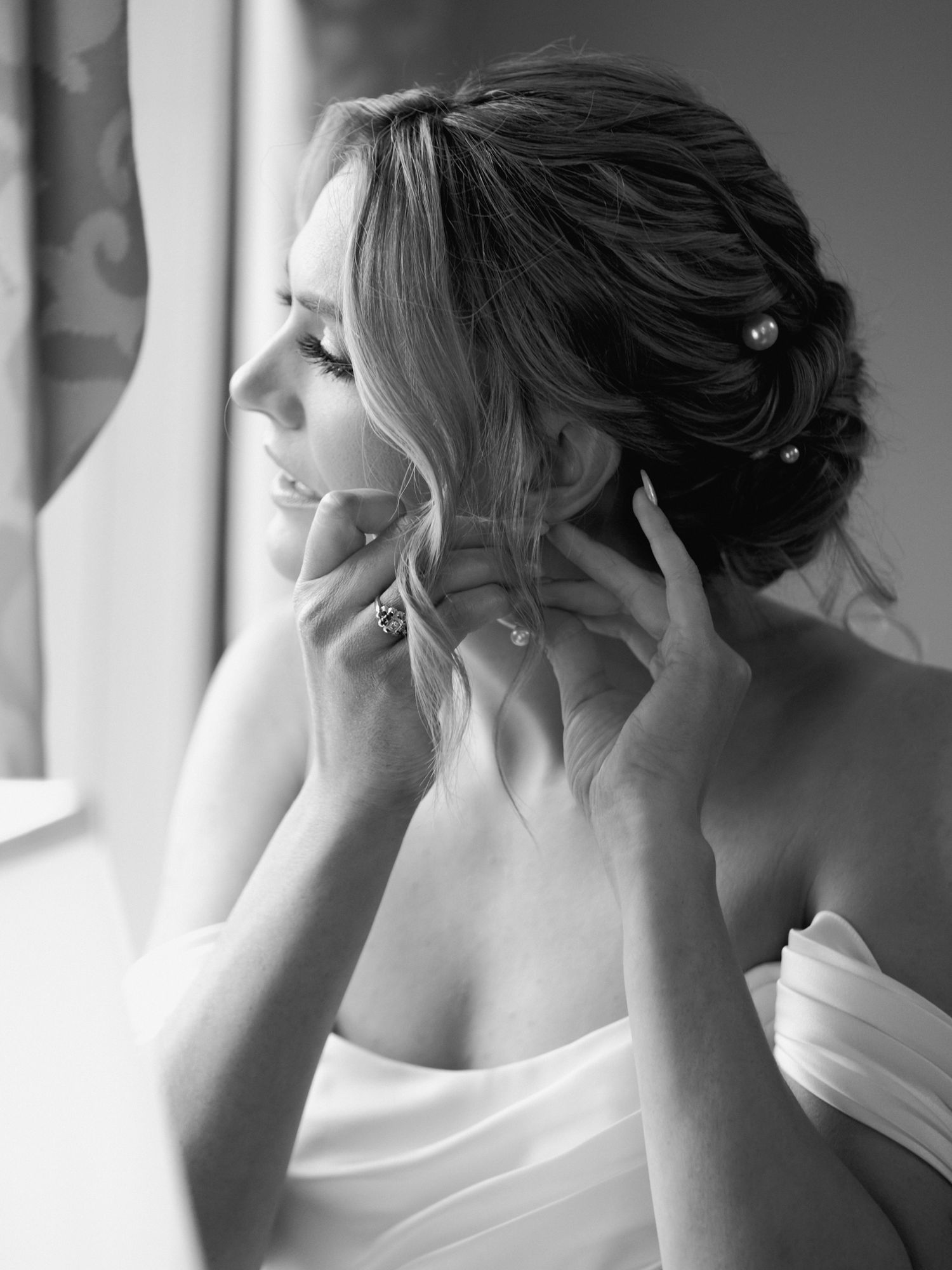 Black and white bridal portrait showing elegant updo hairstyle with soft curls framing the profile near a window.