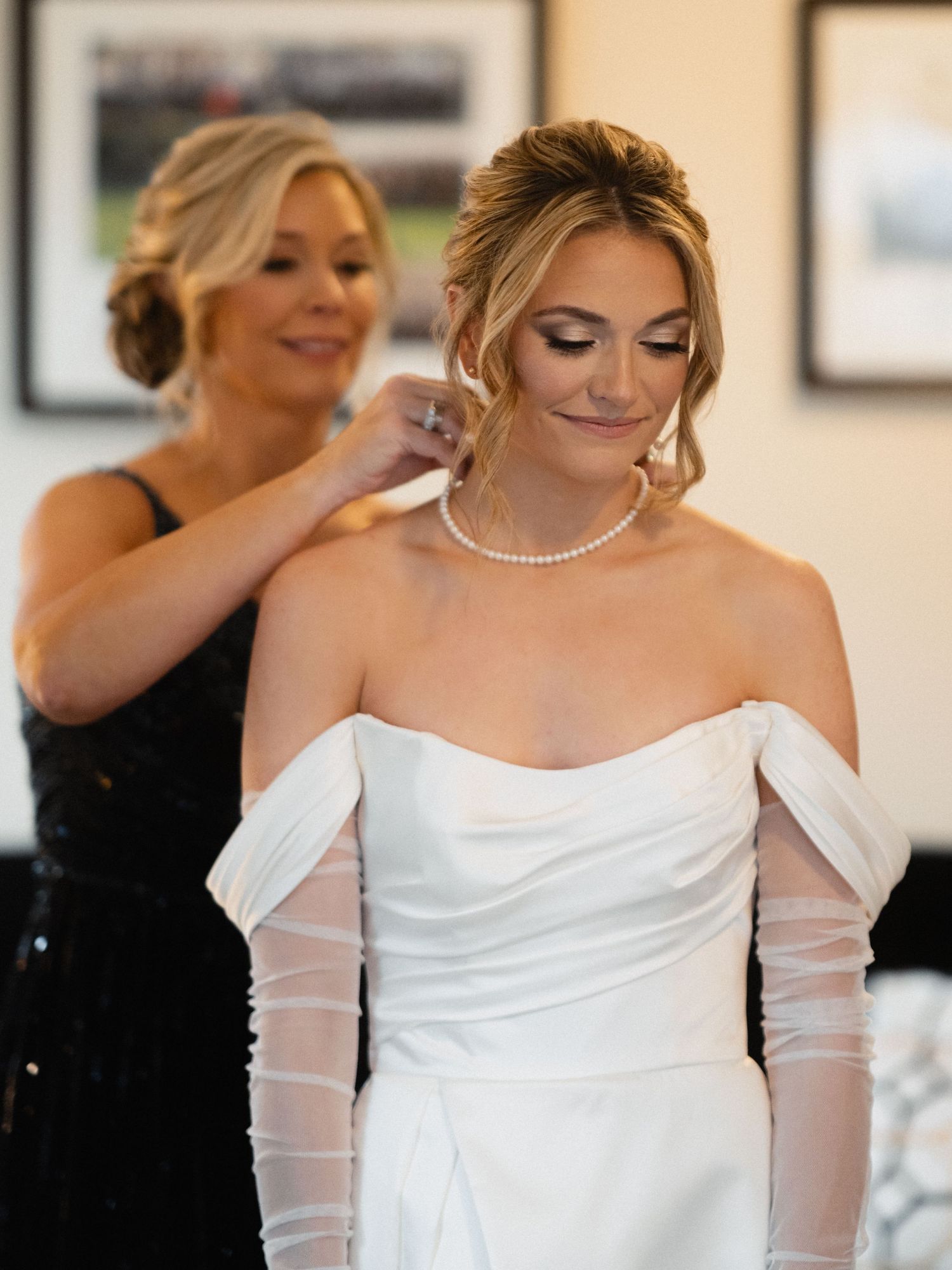 Wedding preparations showing bride in elegant white off-shoulder gown with sheer sleeves and pearl necklace.