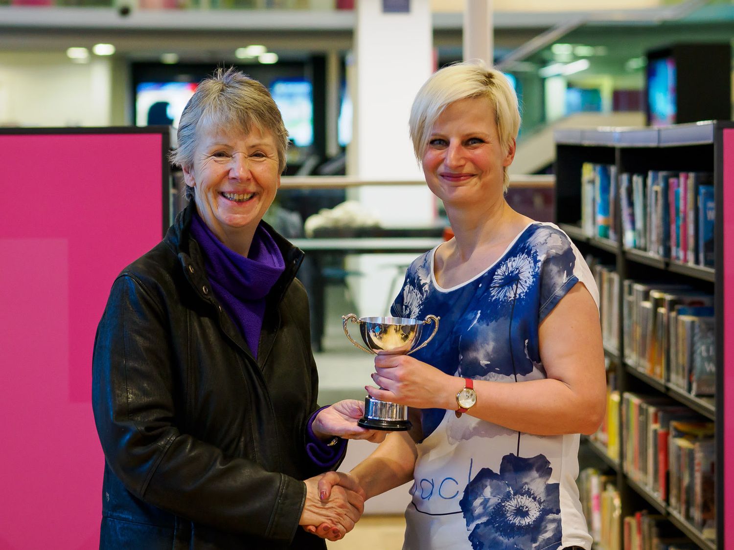 Two women share a handshake and trophy in a library setting.