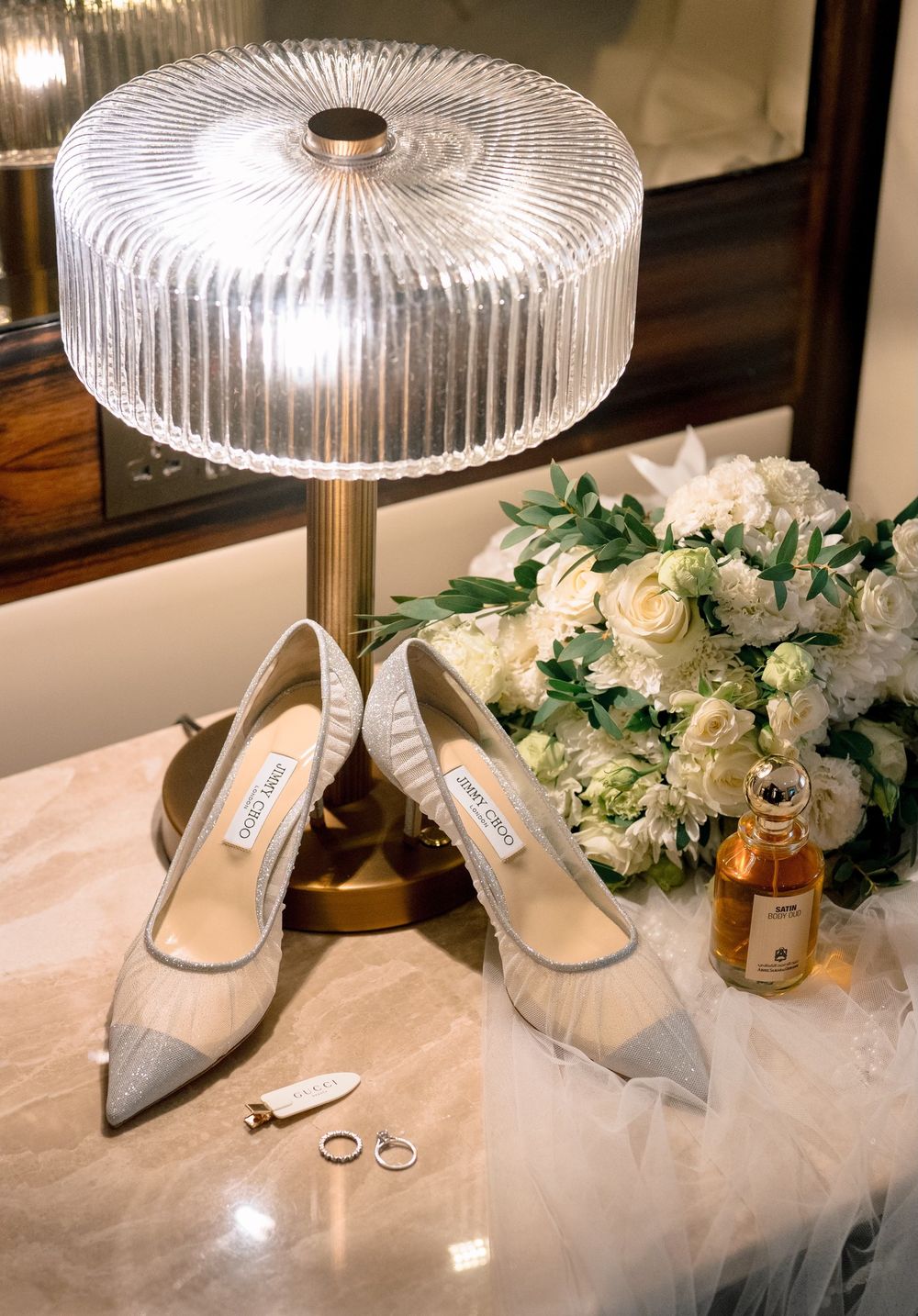 Jimmy Choo bridal heels displayed beside white floral bouquet, gold rings, perfume, and elegant glass lamp on marble surface.