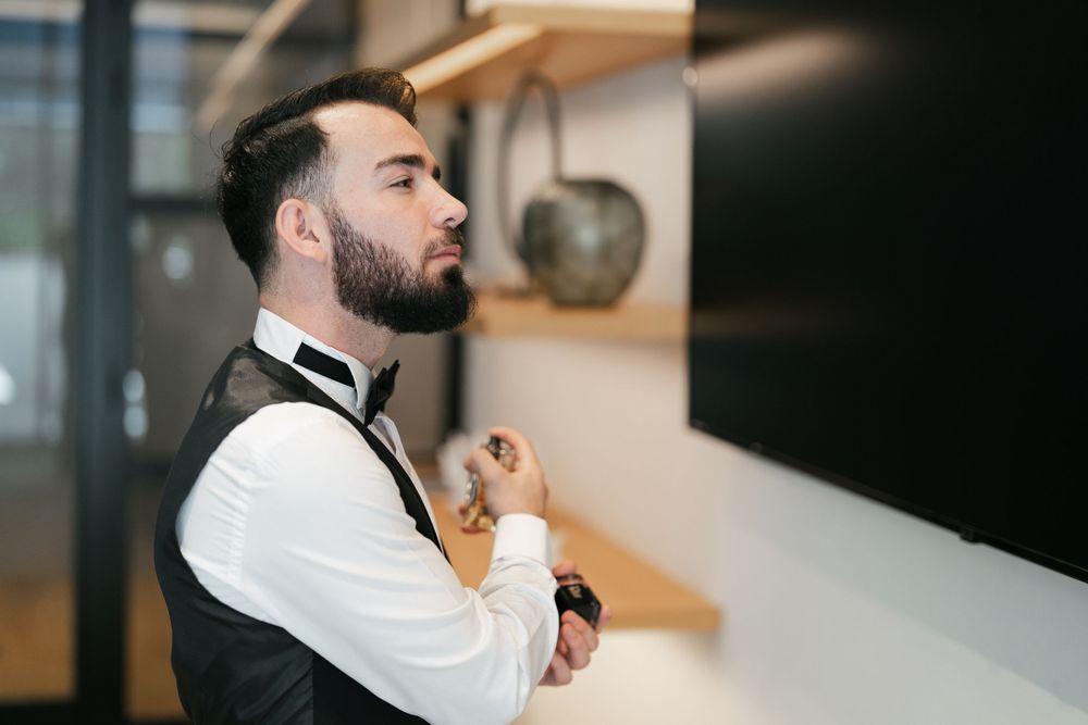 Side profile of a person in formal attire adjusting their black bowtie while standing in a room.