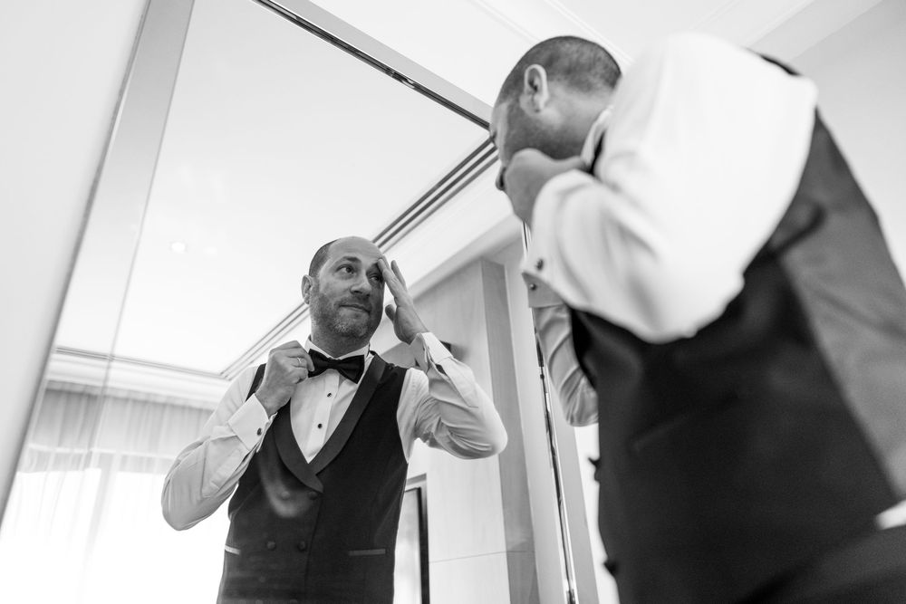 Black and white photo of a groom adjusting his bow tie while looking in a mirror before his wedding.