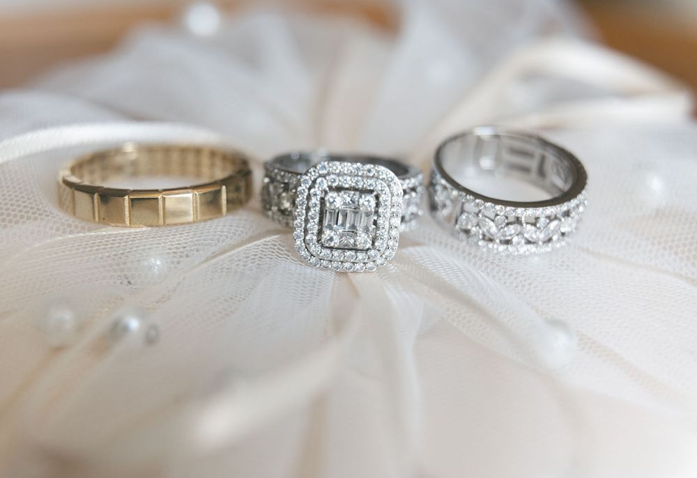 Three wedding rings displayed on white fabric, featuring a diamond engagement ring with double halo setting and two band rings.