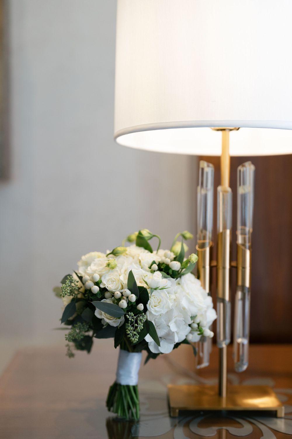 An elegant white floral bouquet with greenery sits beside a modern table lamp with crystal accents and white shade.