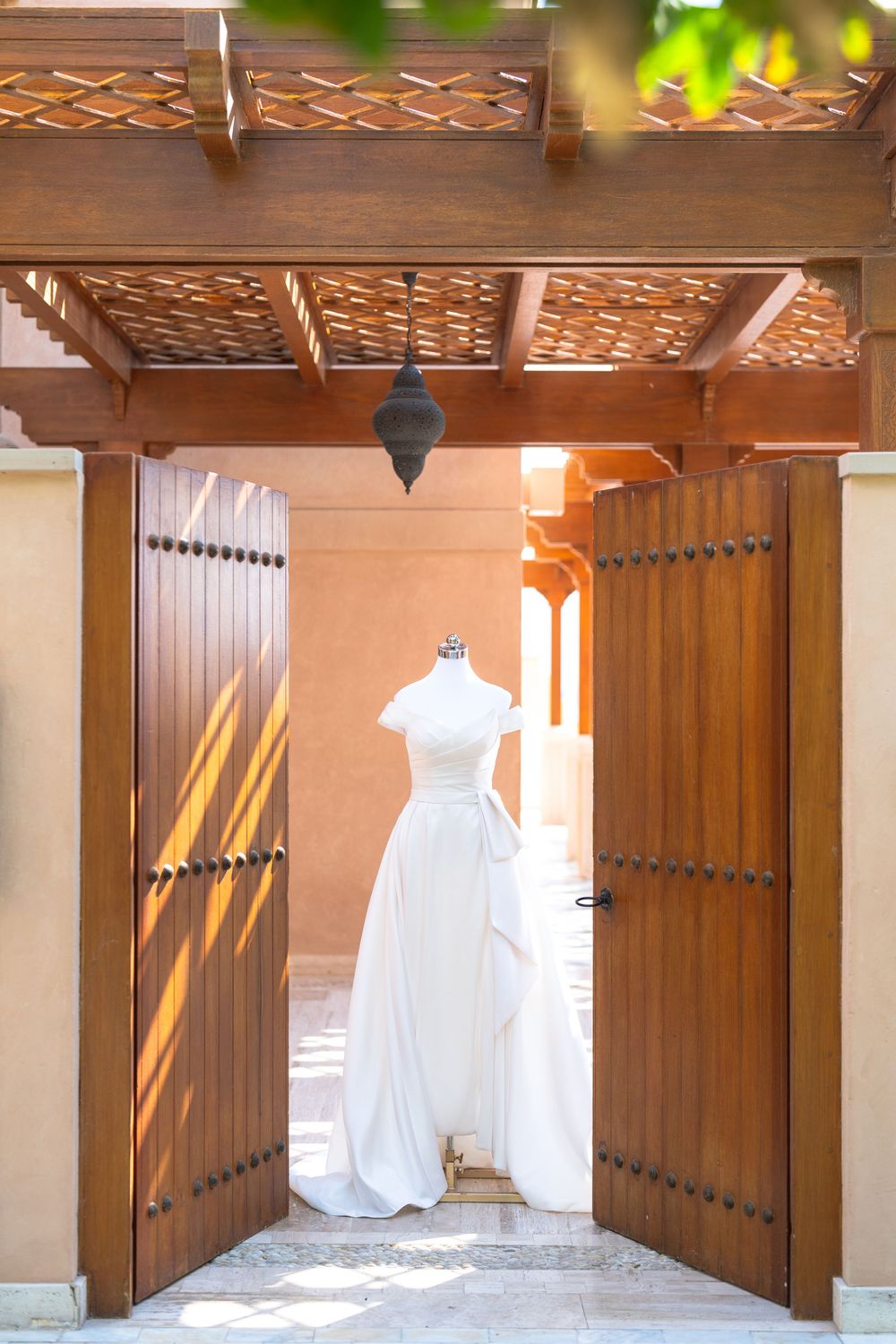 Elegant white wedding dress displayed in wooden doorway with ornate carved pergola overhead casting dramatic shadows.