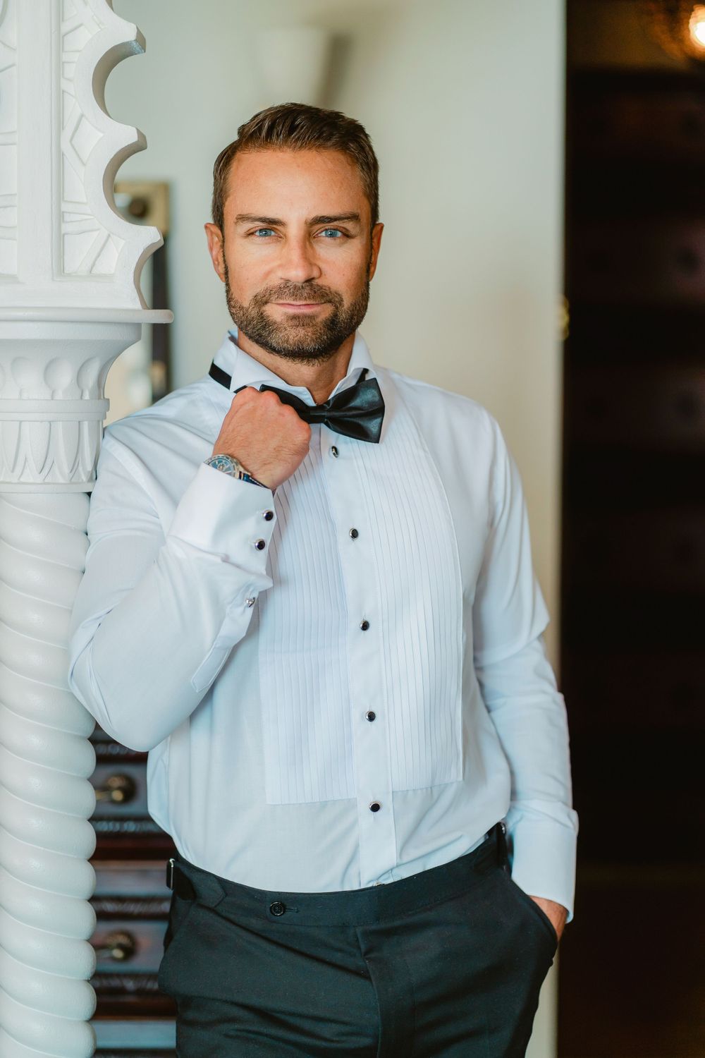 Man in white dress shirt and bow tie posing against ornate white pillar.