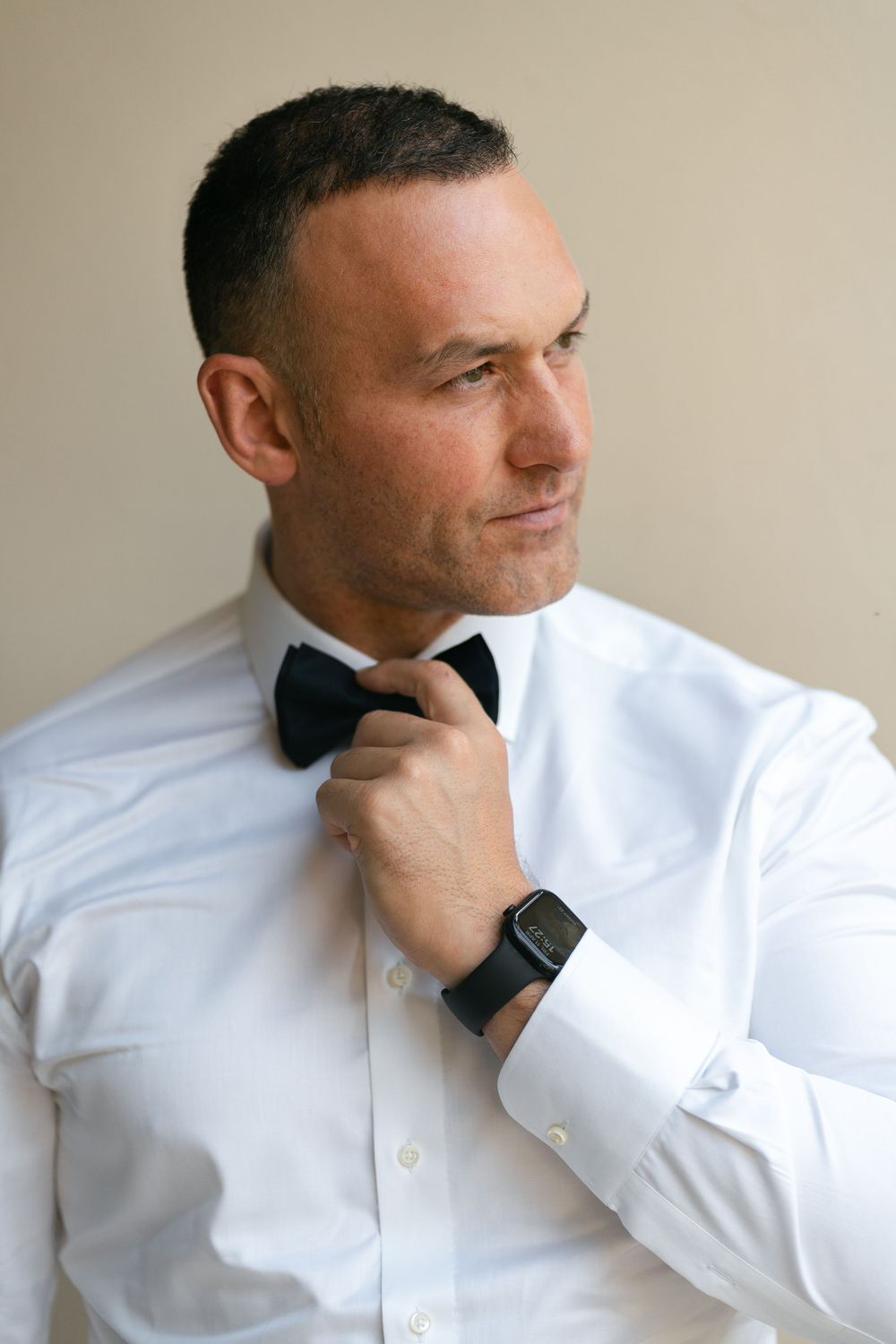 Man in white dress shirt and black bow tie adjusts his collar while wearing a smart watch and looking to the side.