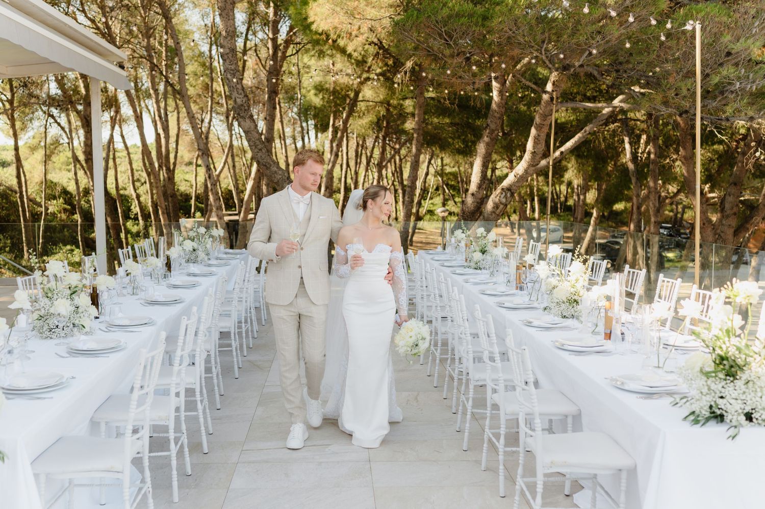 Elegant outdoor wedding reception setup with white chairs and floral arrangements in a wooded area.