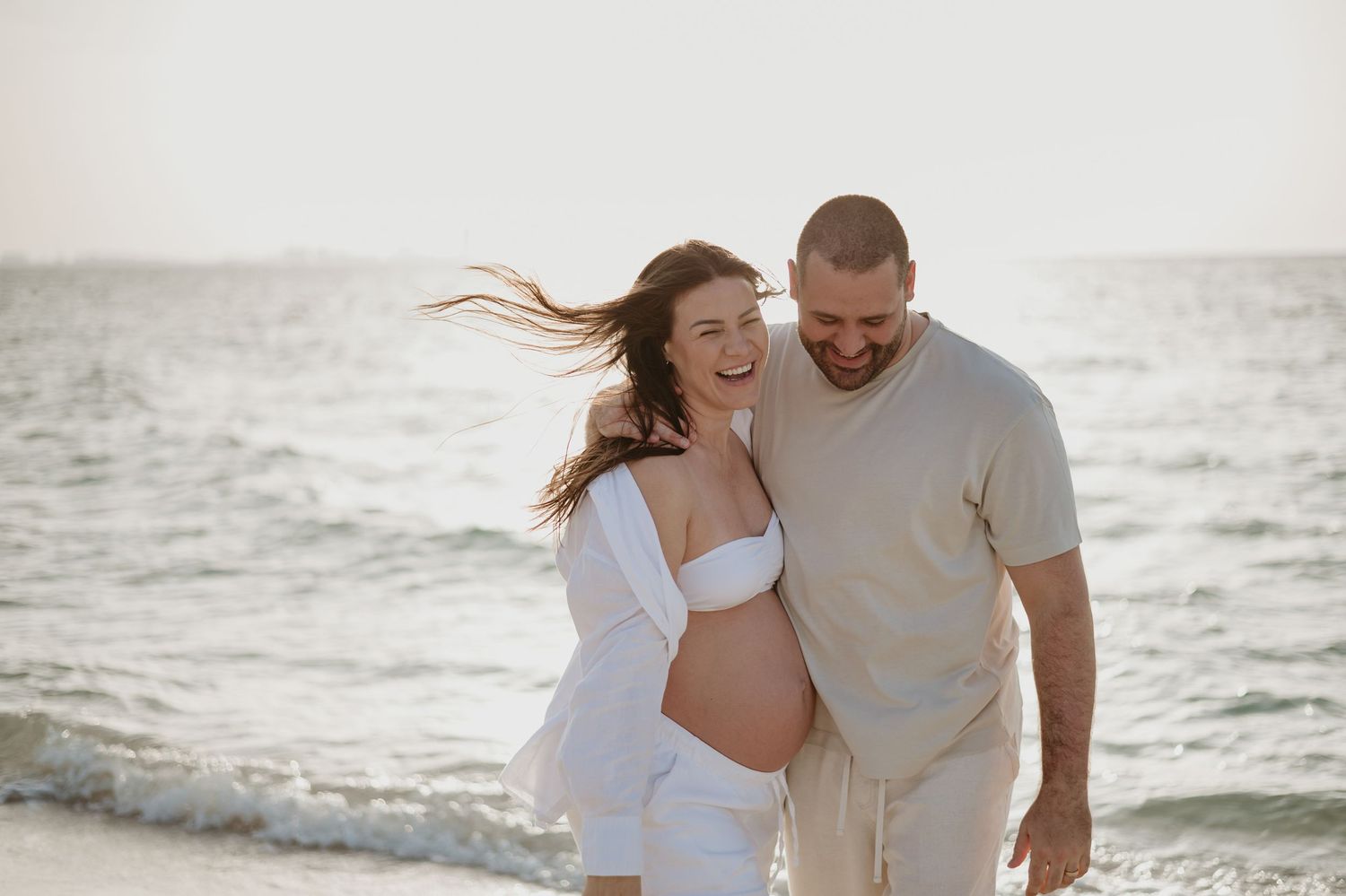 Expectant mother in white dress and sun hat stands peacefully by the ocean at sunset.