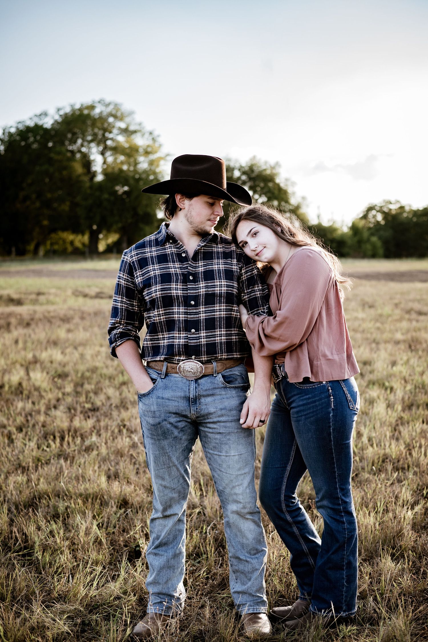 A couple embraces in a grassy field at sunset wearing plaid and denim.