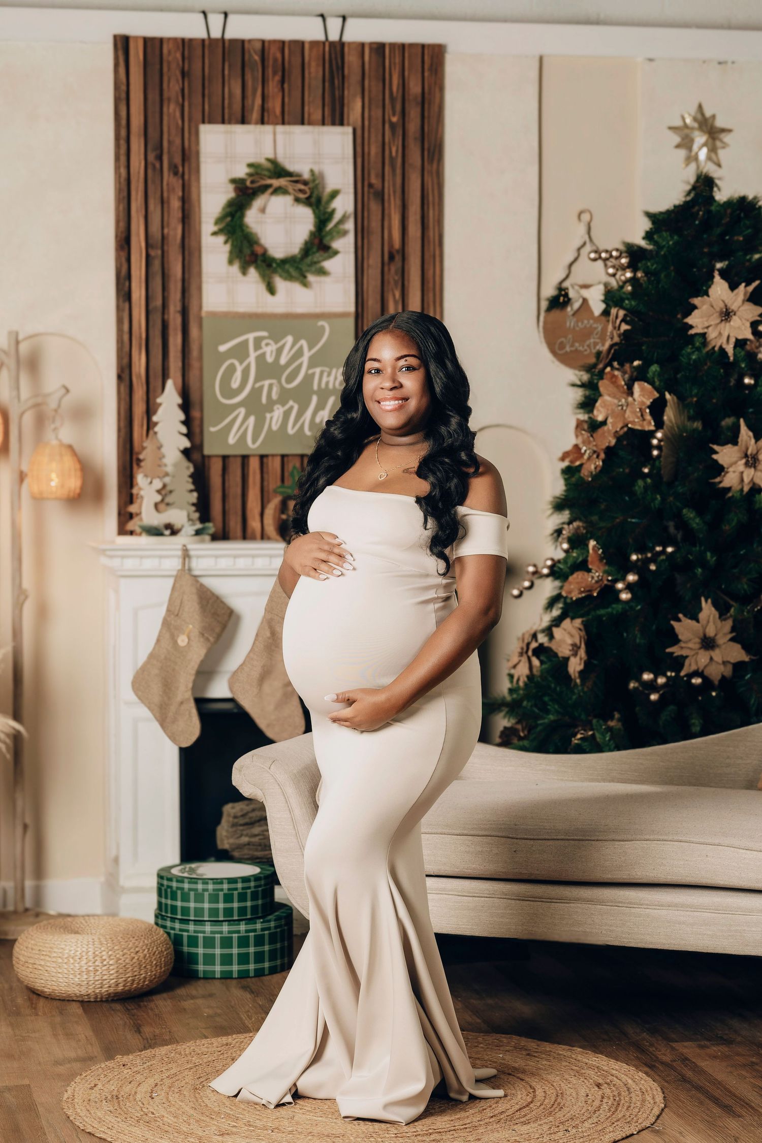 Pregnant woman in elegant white off-shoulder gown posing by Christmas tree and fireplace.