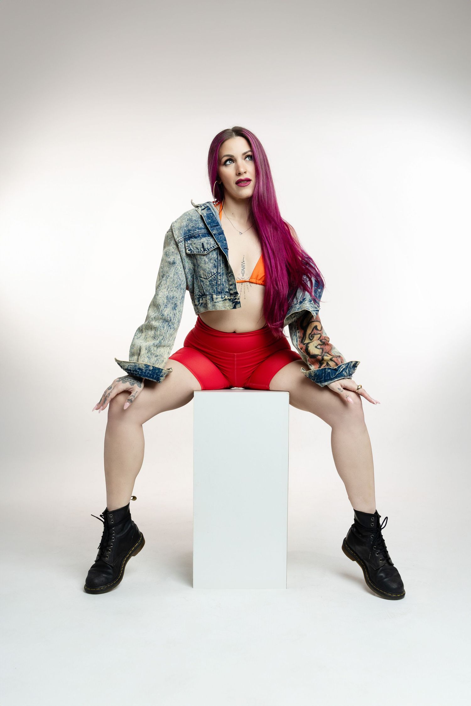 A model in red shorts and cropped denim jacket poses on white cube against plain background wearing combat boots.