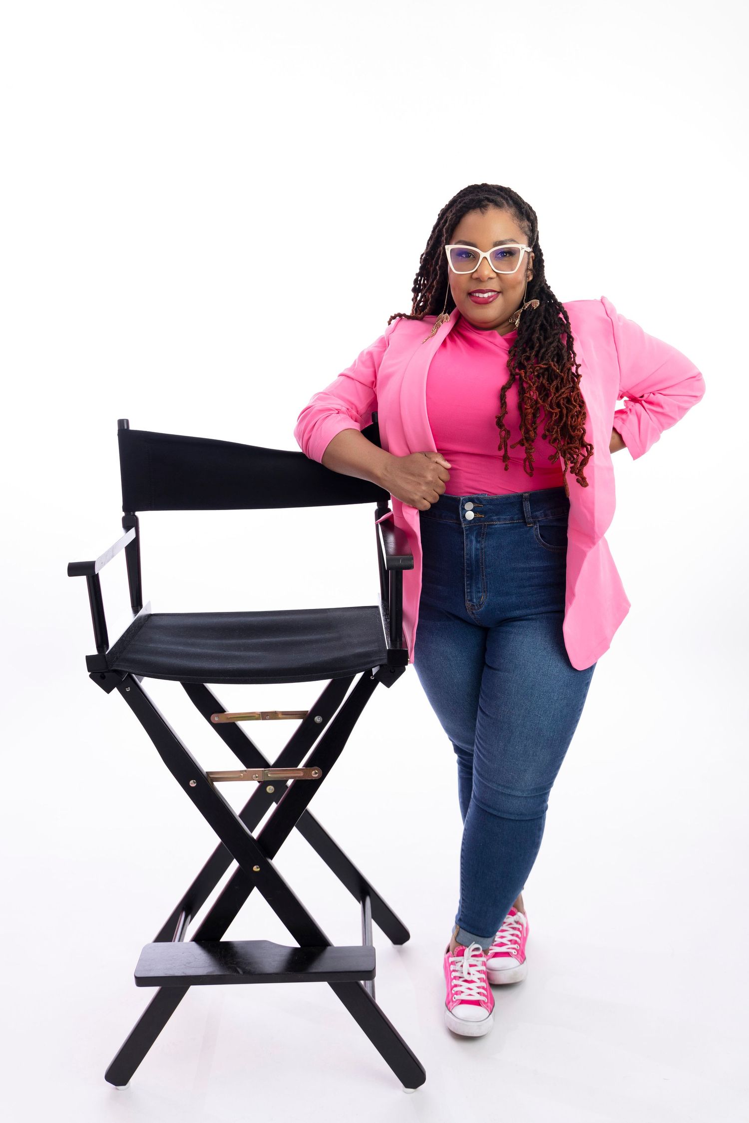 Woman in pink blazer and jeans poses confidently next to director's chair.
