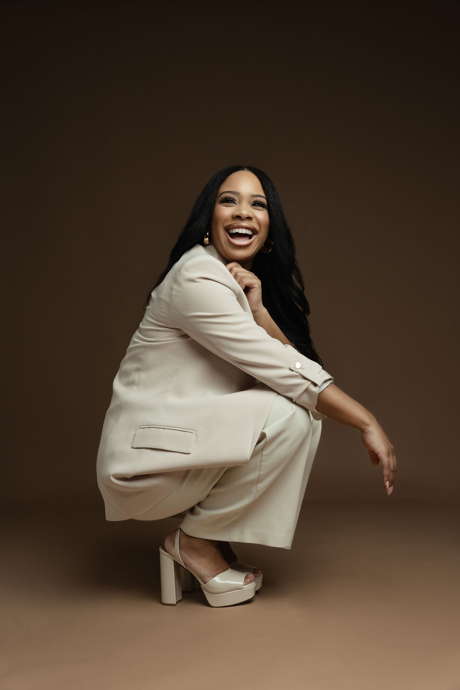 A person in a cream-colored pantsuit and platform shoes crouches with a joyful expression against a brown backdrop.