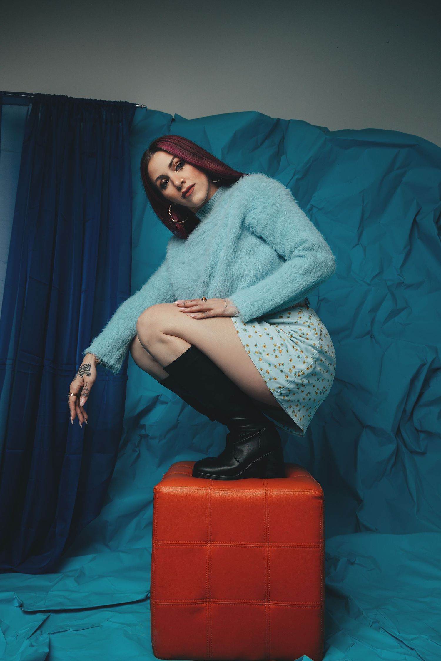Fashion portrait with fuzzy blue sweater and lace dress seated on red ottoman against turquoise backdrop.
