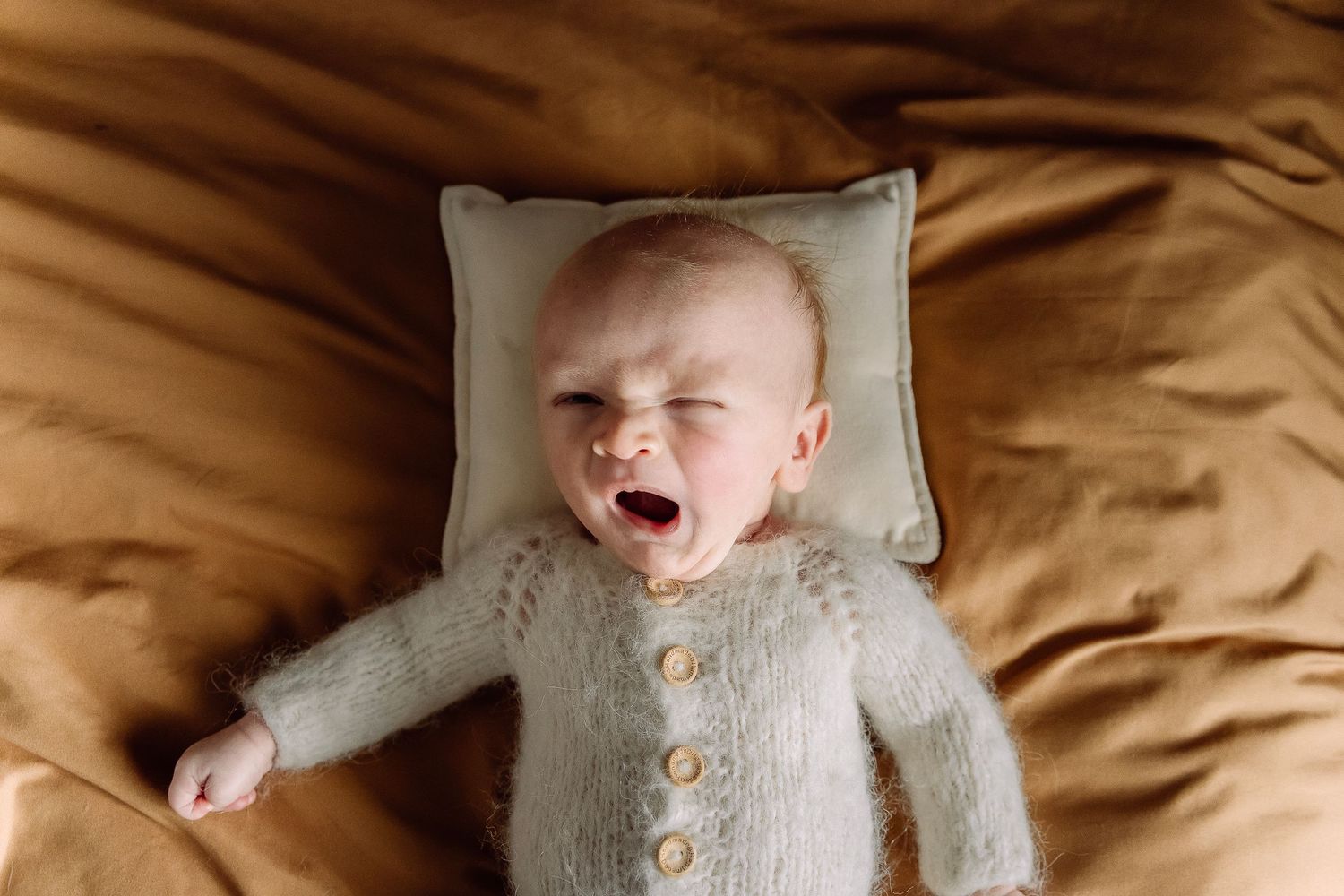 Baby in gray knit cardigan lying on beige pillow with expressive yawning expression.