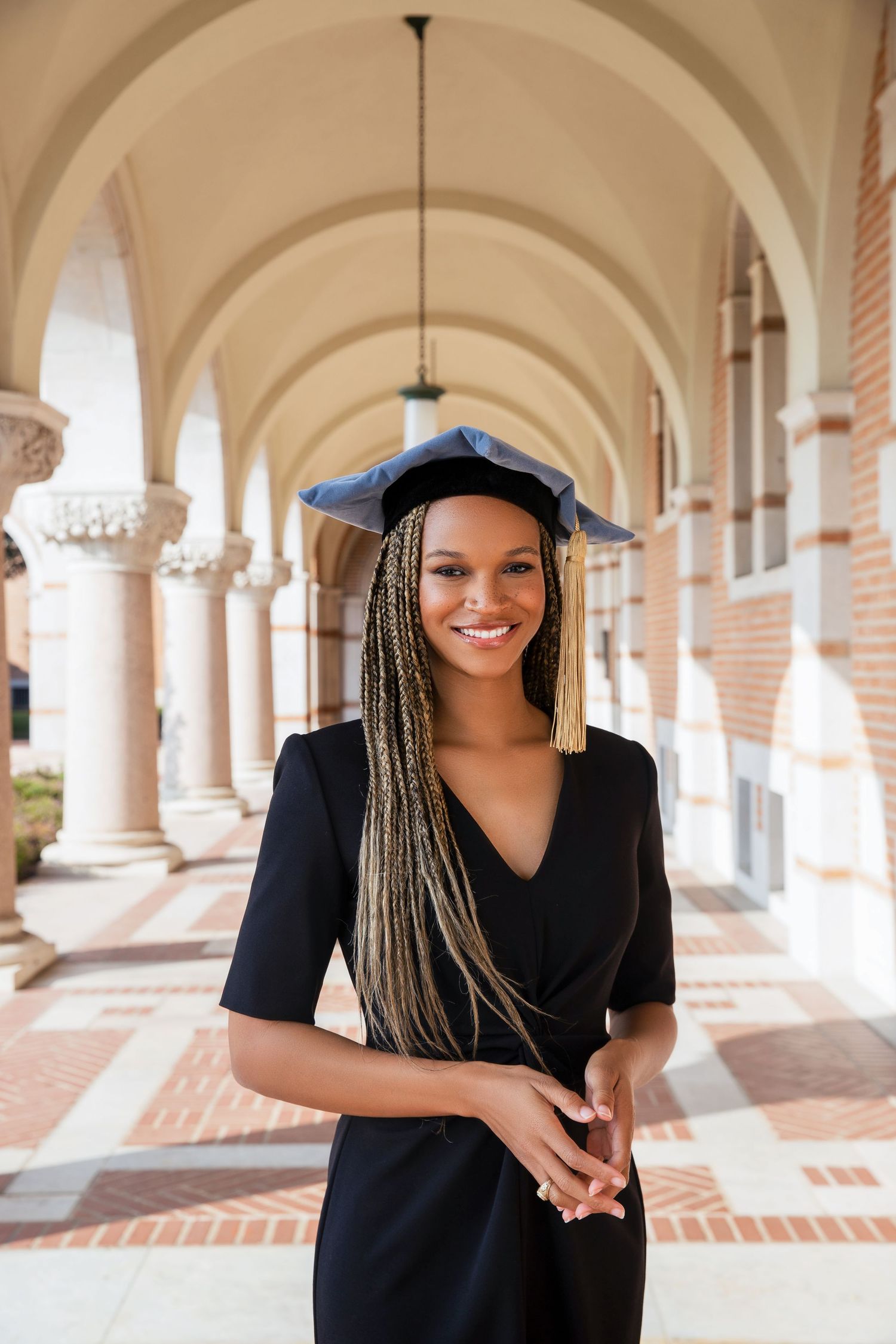 Graduate dressed in black academic regalia with long braids stands smiling in an arched university colonnade.