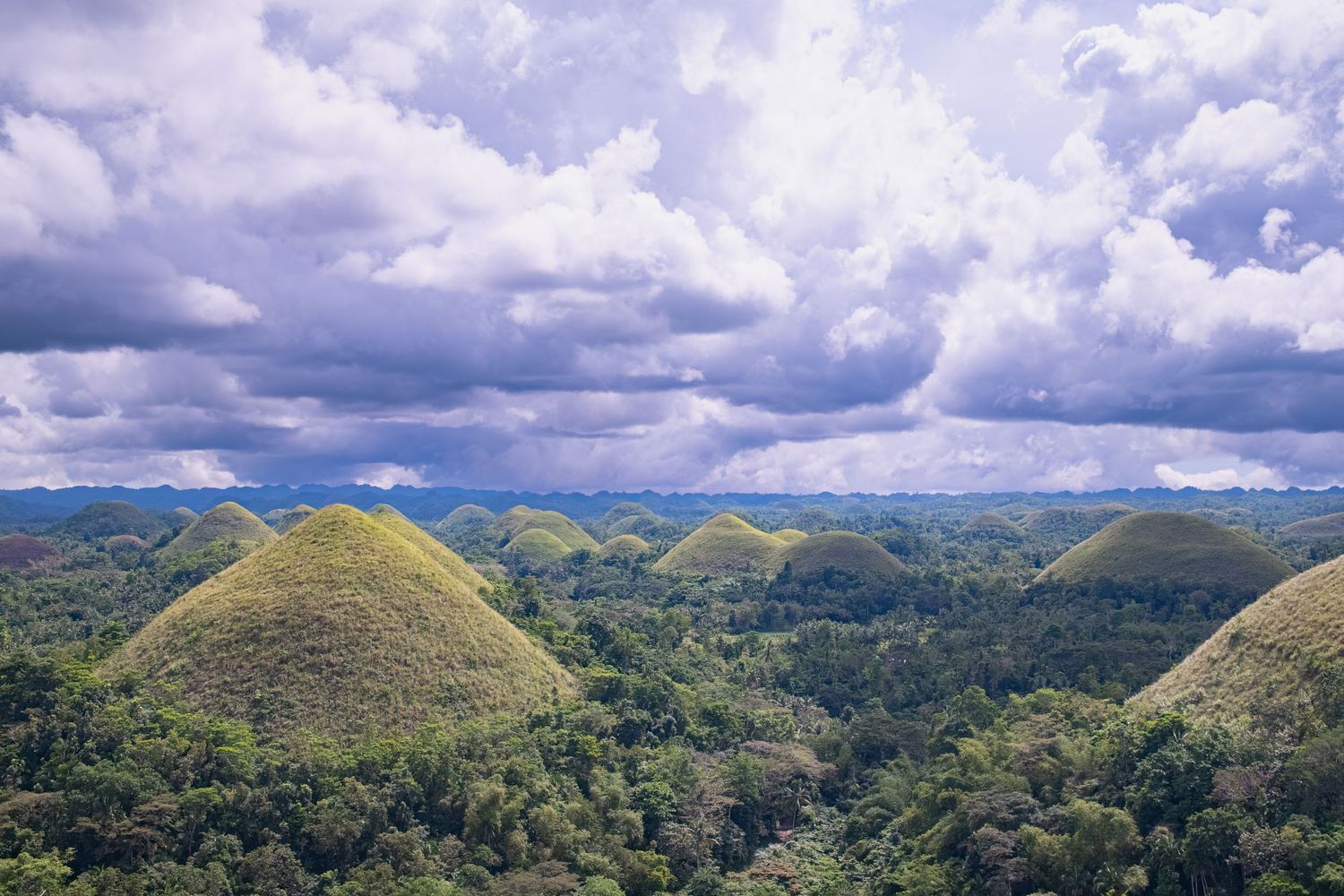 Rolling green hills and mountains under cloudy skies in a tropical landscape photographed by Gia Quilap.