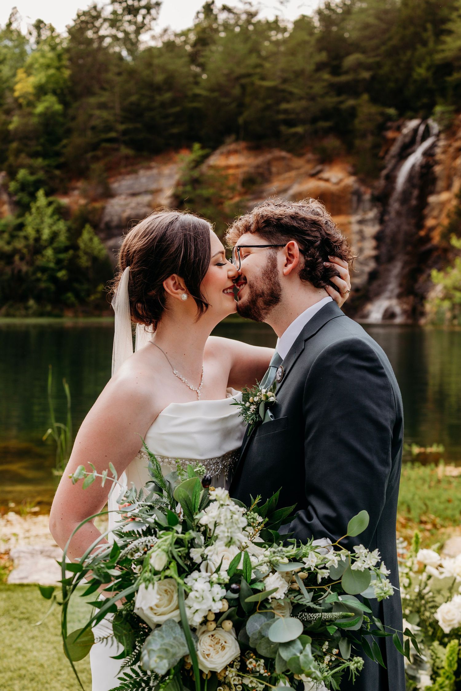 Bride and groom share romantic kiss with lush waterfall and lake backdrop on wedding day.