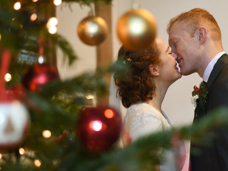 A bride and groom share a kiss in front of a beautifully decorated Christmas tree at St Dunstan’s Church. Photography by Abigail's Studio.