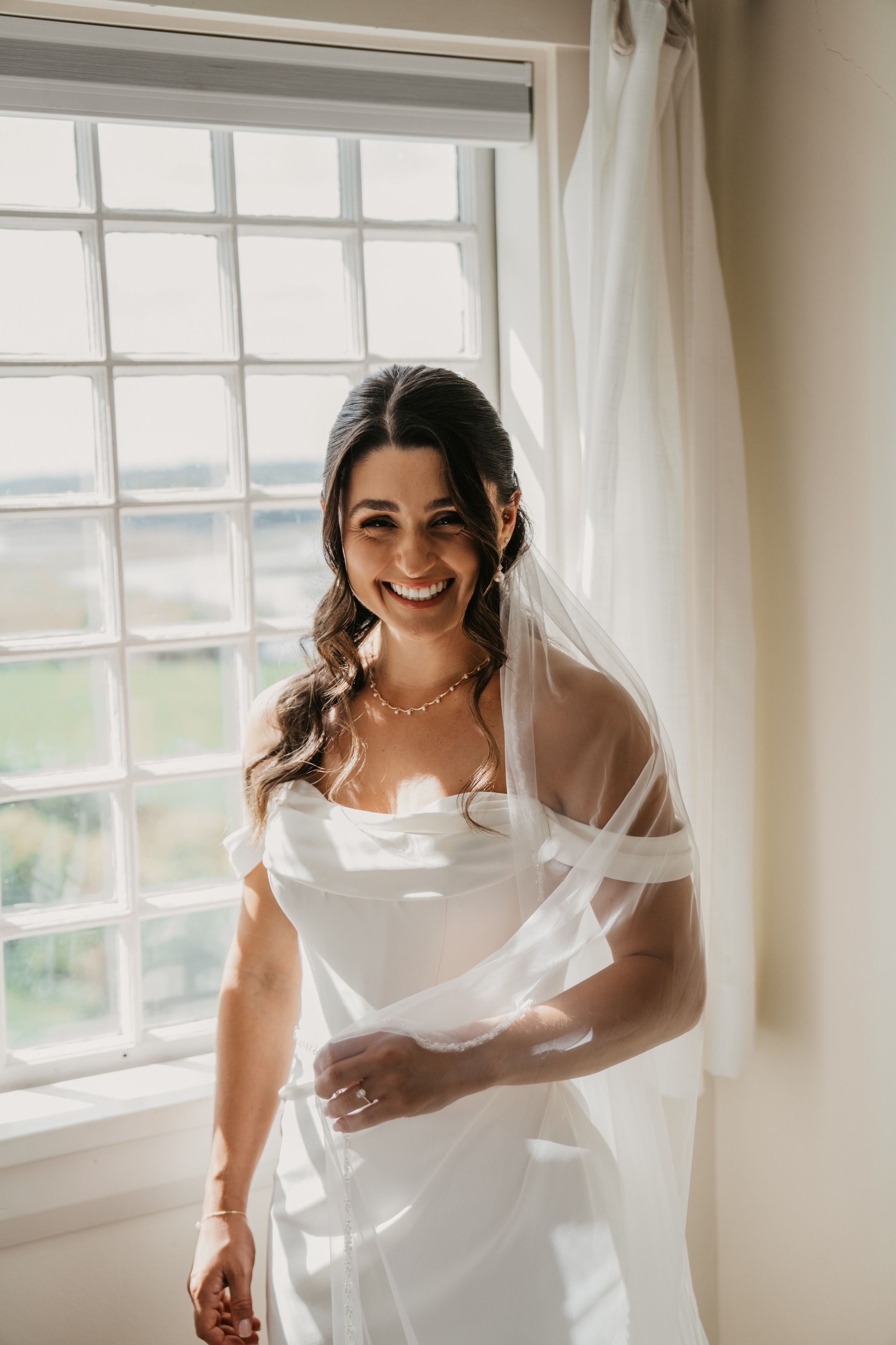 A bride in a white wedding dress and veil stands by a sunny window with sheer curtains.