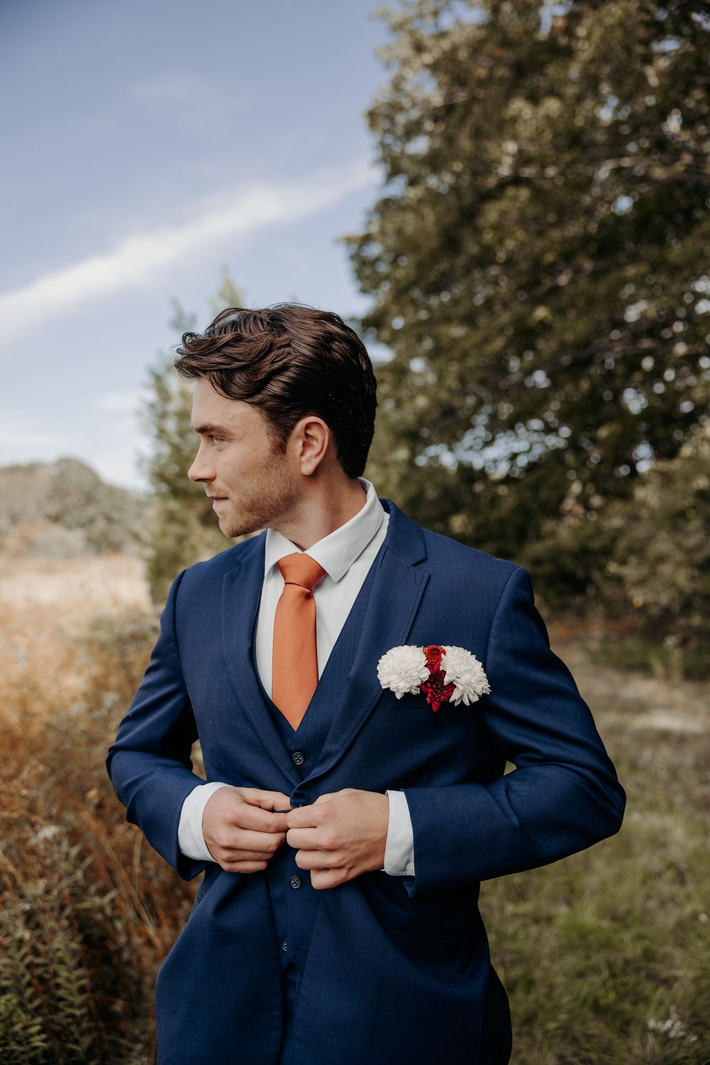 A groom in a navy blue suit with orange tie adjusts his jacket while standing outdoors on his wedding day.