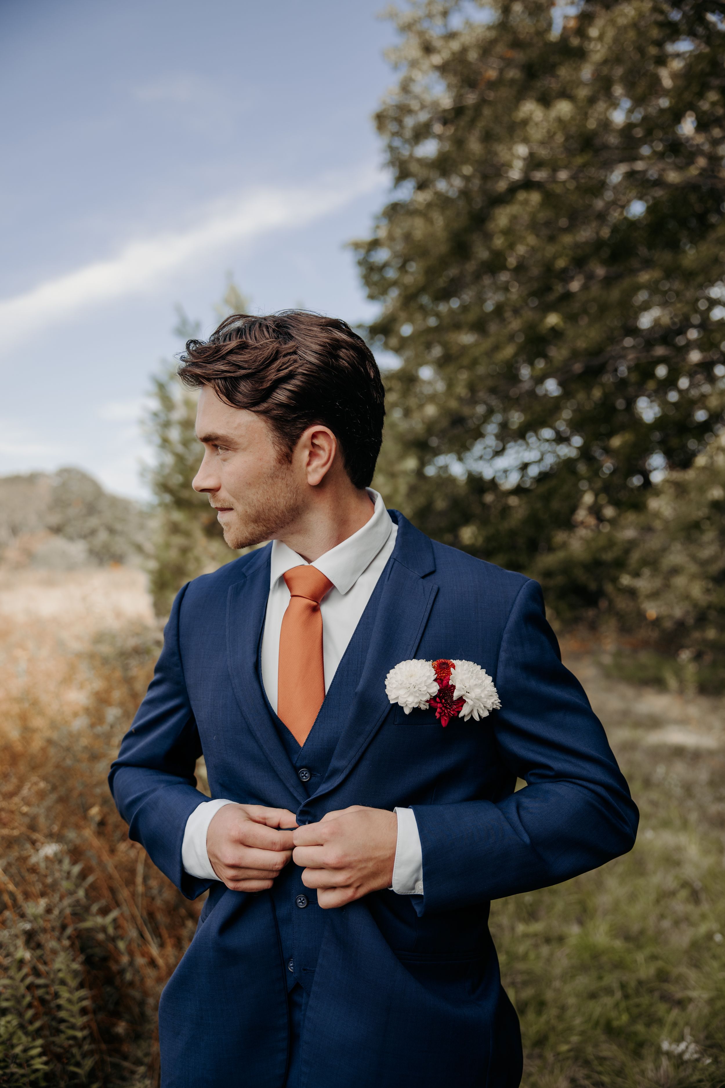 A groom in a navy blue suit with orange tie adjusts his jacket while standing outdoors on his wedding day.