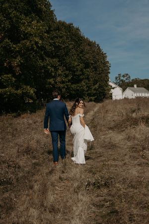 A couple in wedding attire walking up a grassy hill with trees and a white barn in the background.