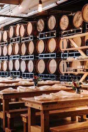 Rustic wooden tables and benches arranged in front of a wall of aging whiskey barrels in a distillery tasting room.