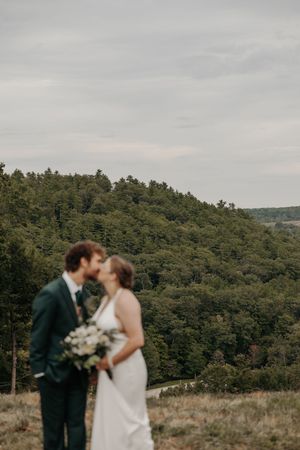 A couple shares a romantic moment during their wedding photoshoot on a hilltop overlooking a forested mountain landscape.