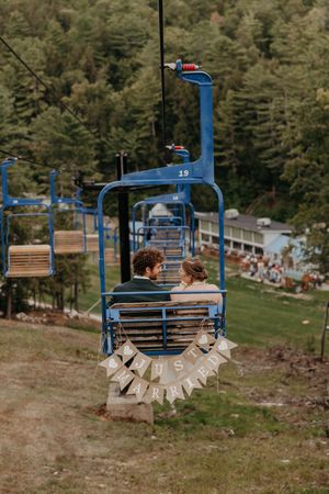 Rustic wedding decor with 'Just Married' banner hanging on blue ski lift chairs against forest backdrop.