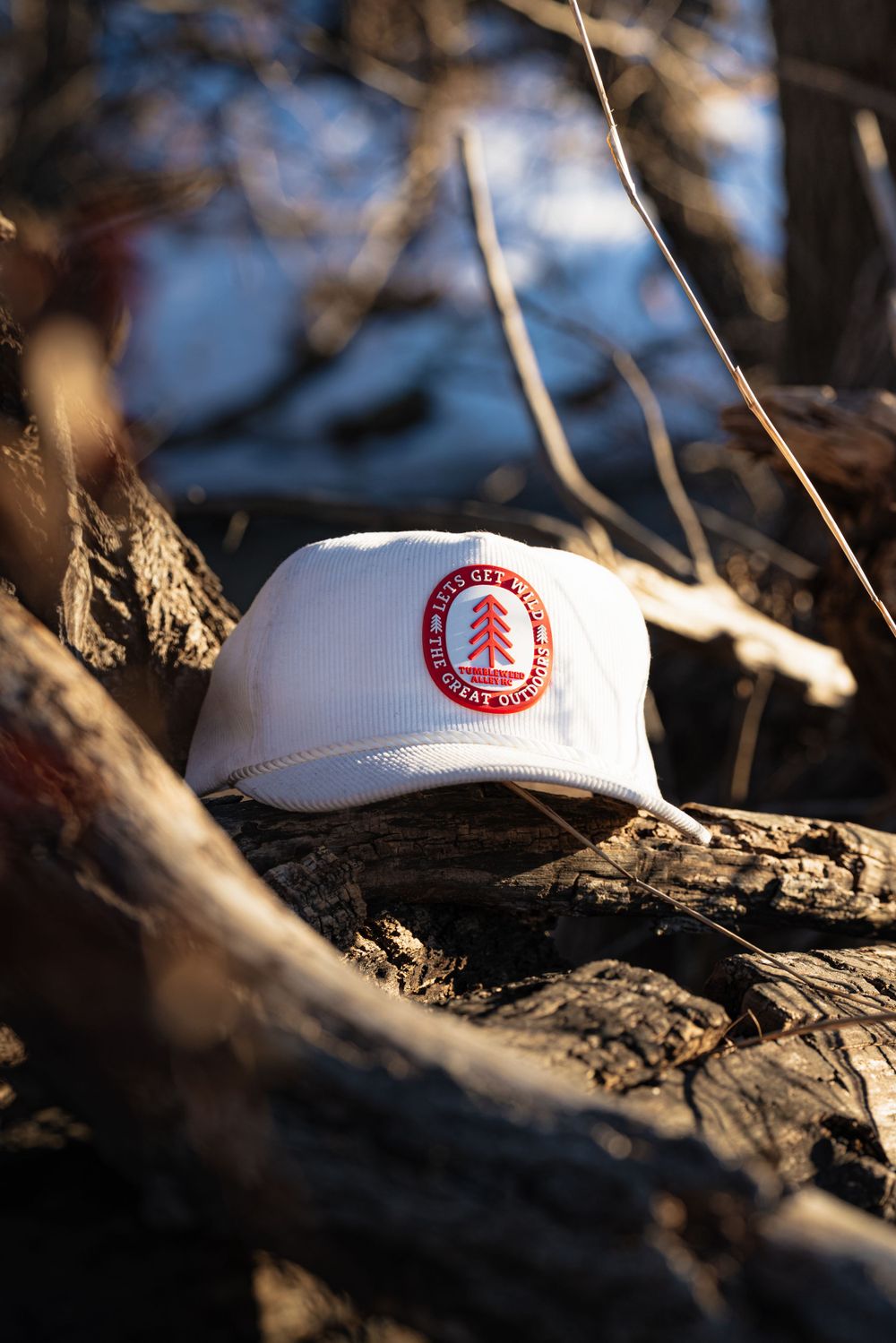 A white baseball cap with a red circular logo featuring a pine tree sits on weathered wood in natural lighting.
