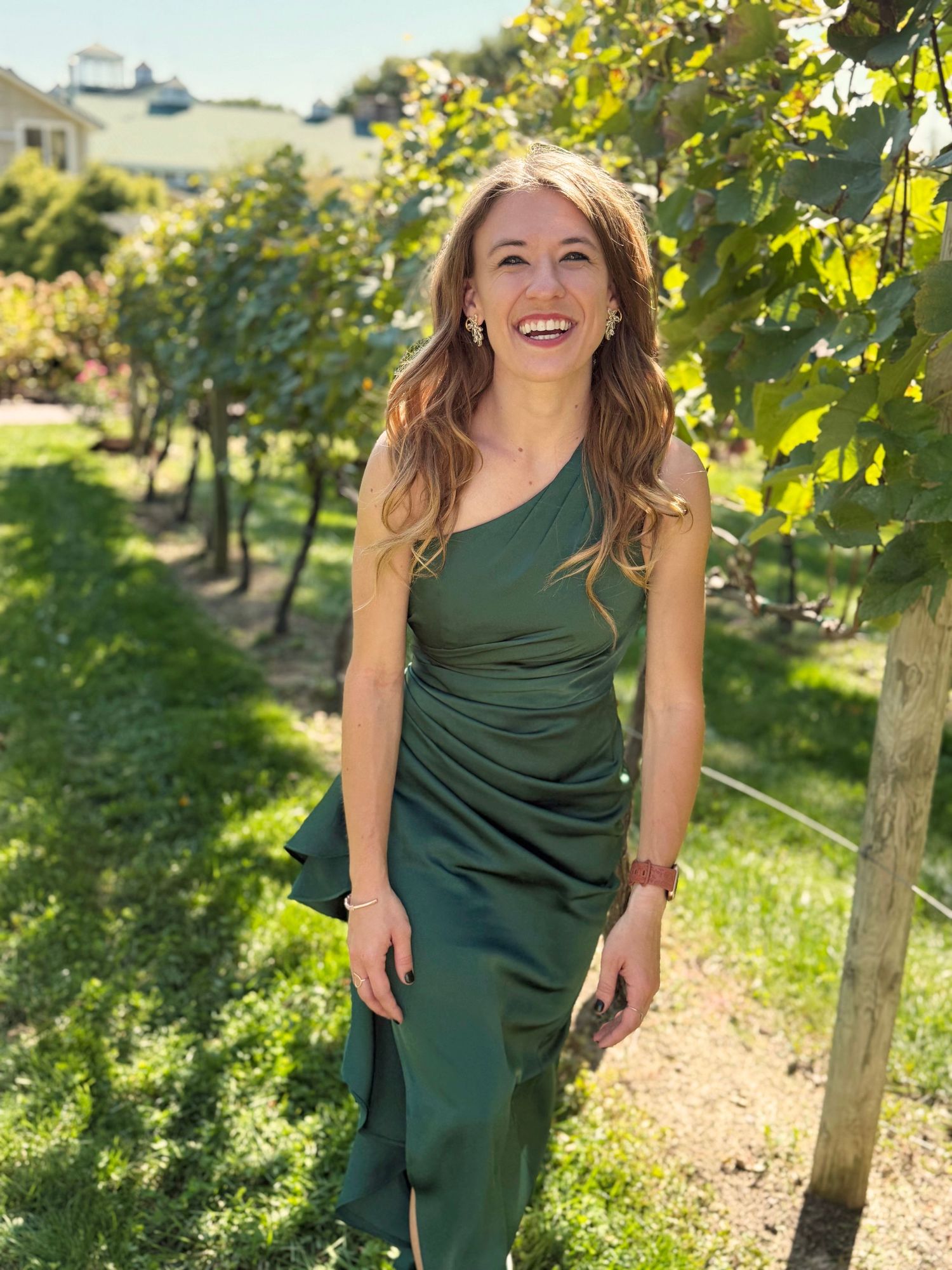 Woman in green dress smiling while standing in a sunny vineyard.