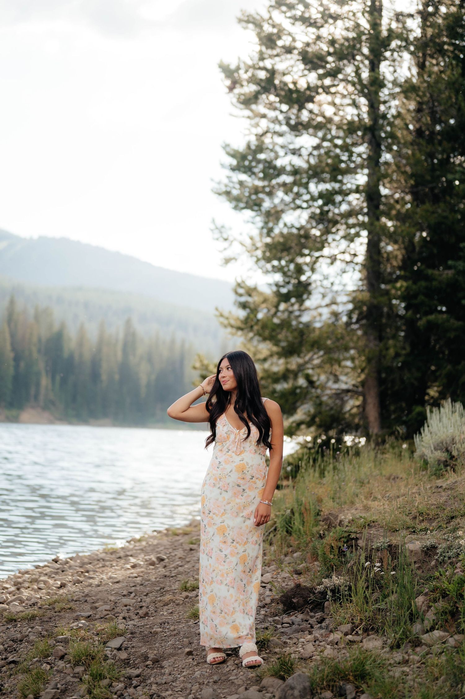 Woman in floral maxi dress standing by a mountain lake surrounded by tall pine trees.