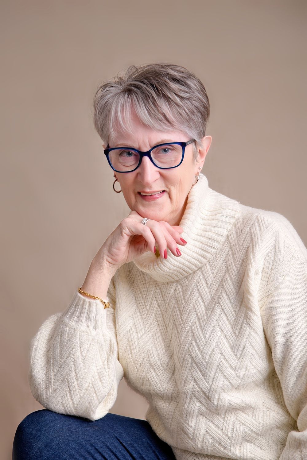Senior wearing glasses and cream turtleneck sweater poses with hand resting on chin against neutral background.