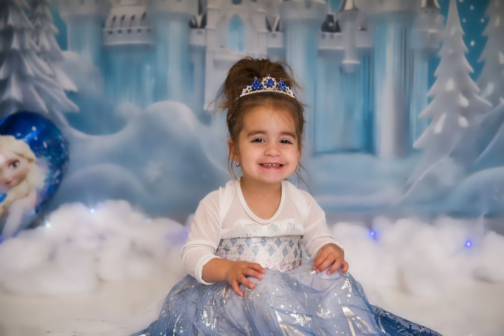A child in a sparkly blue and white princess dress and tiara poses in a magical winter wonderland setting with snowy decor.