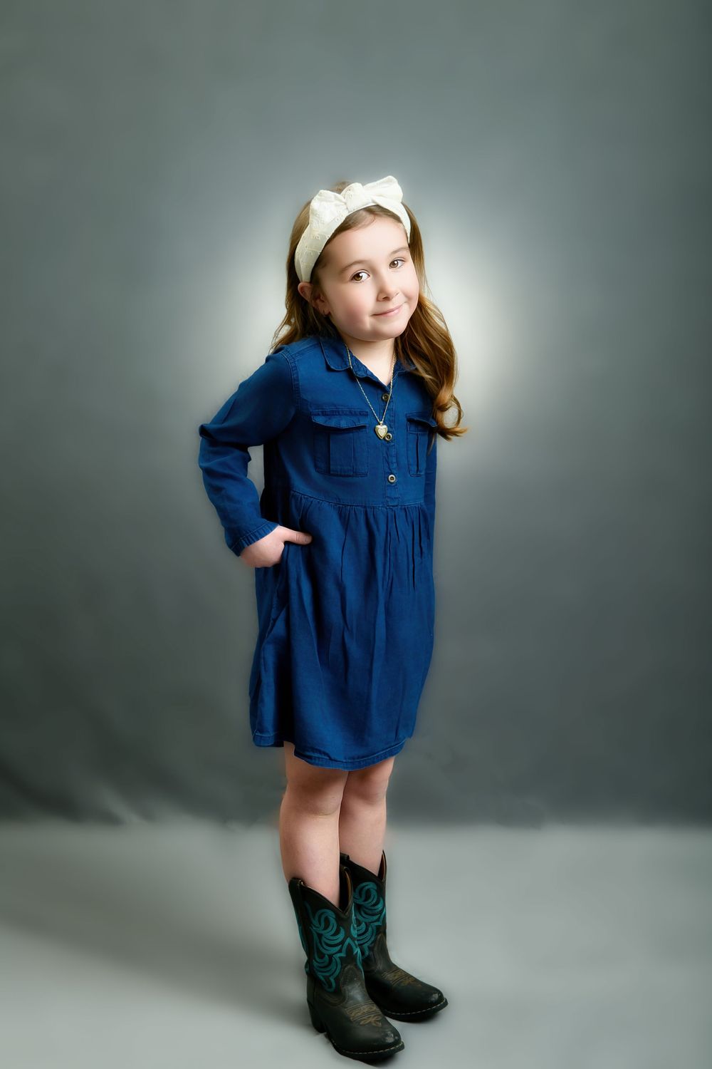 Young child in navy blue dress poses playfully in cowboy boots against grey studio backdrop in series of candid shots.