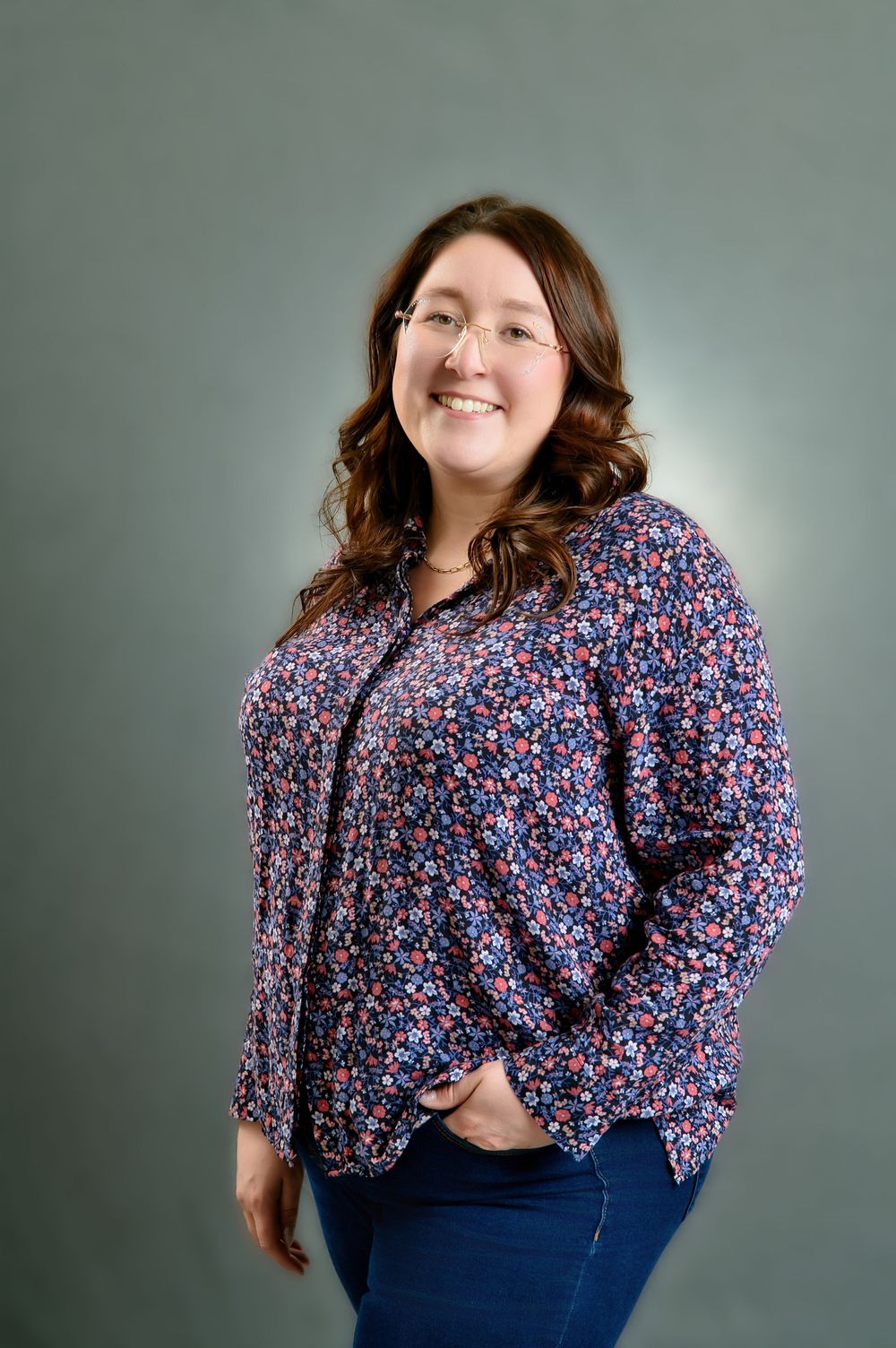 A person wearing a floral print blouse and blue jeans poses against a gray studio background.