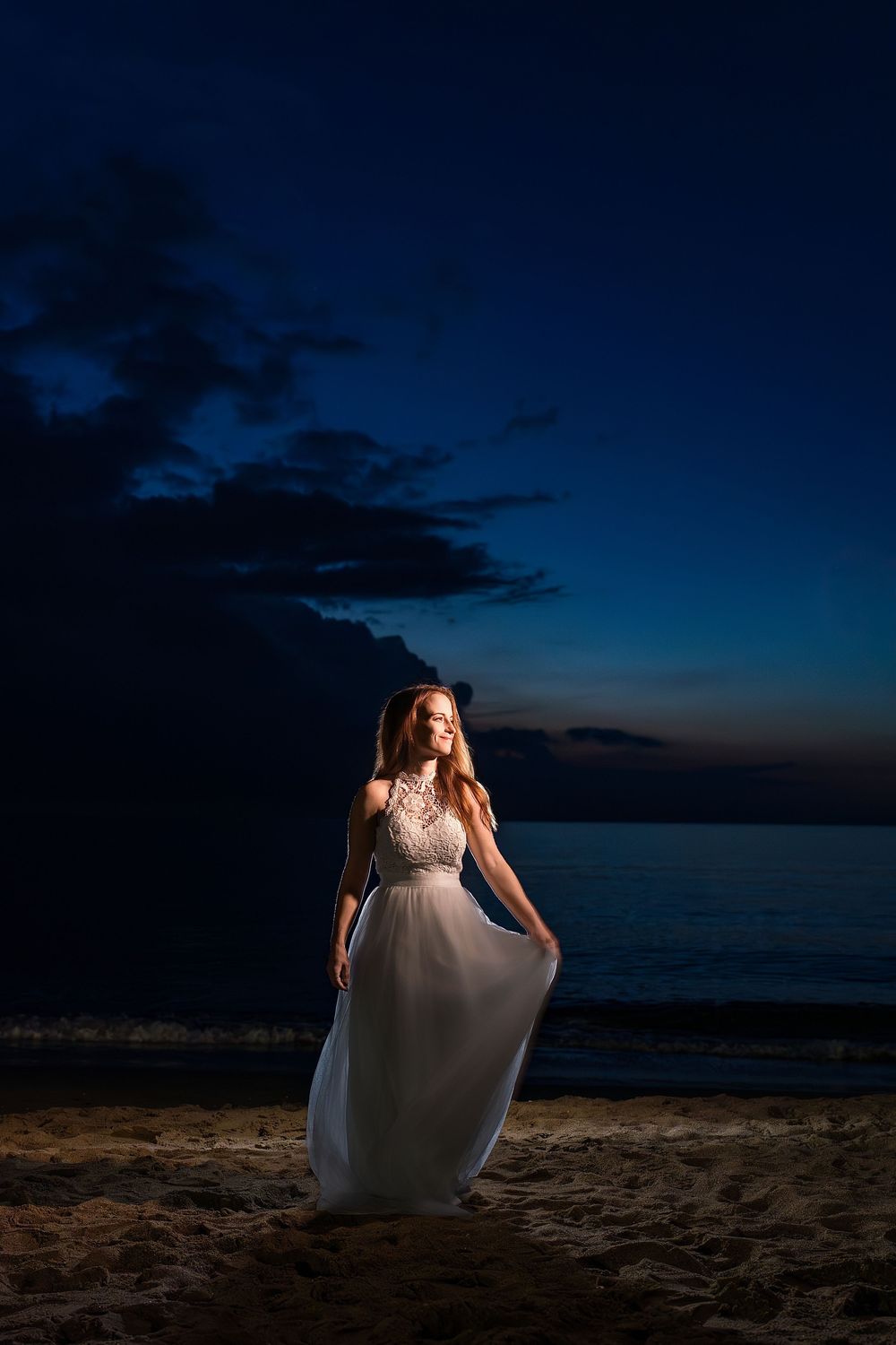 A bride in a white wedding dress dances on a beach at sunset with dramatic dark blue skies in the background.